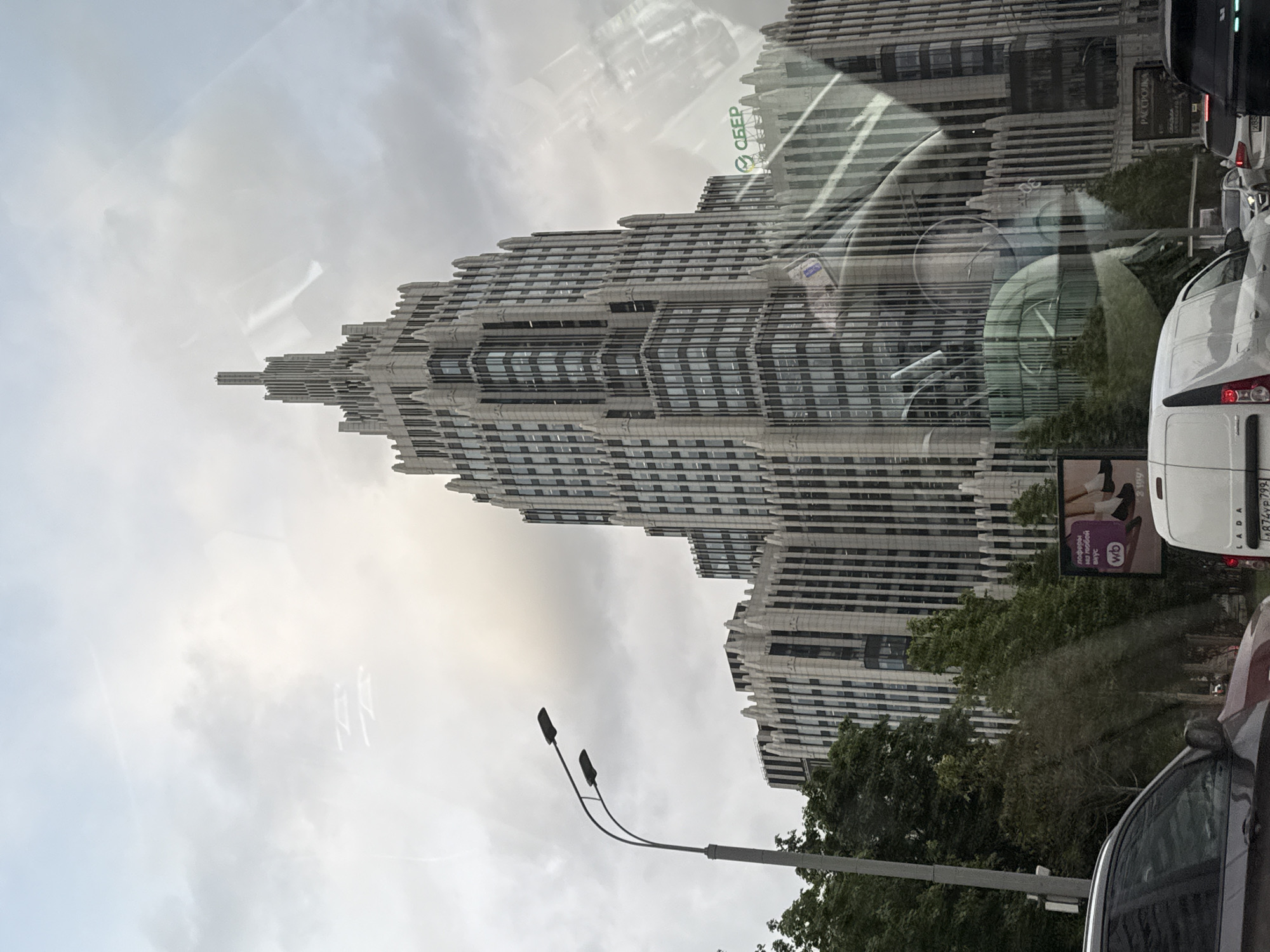 The image shows a tall, light-colored skyscraper with a stepped, tiered design, suggestive of Art Deco architecture. A large "Sber" sign is visible on the building. The image is taken from inside a vehicle, with reflections on the window. Traffic includes a white van with the license plate number "H874YP 197" and the word "LADA" and a commercial sign featuring a footwear image are partially visible in the lower right. The foreground contains part of the street, trees, and streetlights. The sky is overcast with grey clouds. Based on the distinctive architecture and the "Sber" signage, the location is likely near the Sberbank headquarters in Moscow, Russia.