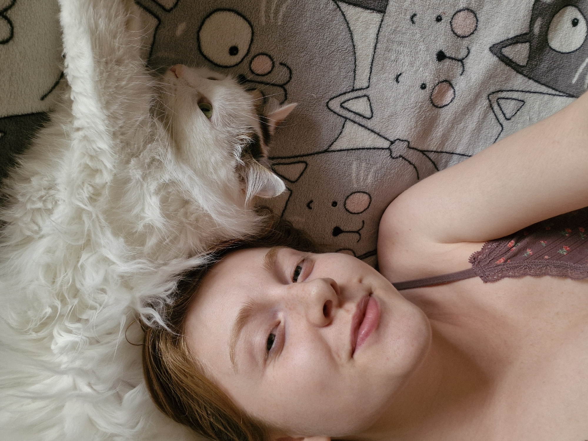 A high-angle, medium close-up photograph shows a woman lying on her back with a white cat on her head. The woman has pale skin, reddish-blonde hair, and is wearing a floral-patterned bralette. She is looking directly at the camera with a relaxed expression. The cat is mostly white with some brown markings on its head and is positioned on the left side of the frame, resting on a soft white fur-like material. Beneath the woman and cat is a gray blanket with a cartoon cat pattern. The setting is indoors, likely a home.
There is no information that this image was taken in Yekaterinburg, Russia.