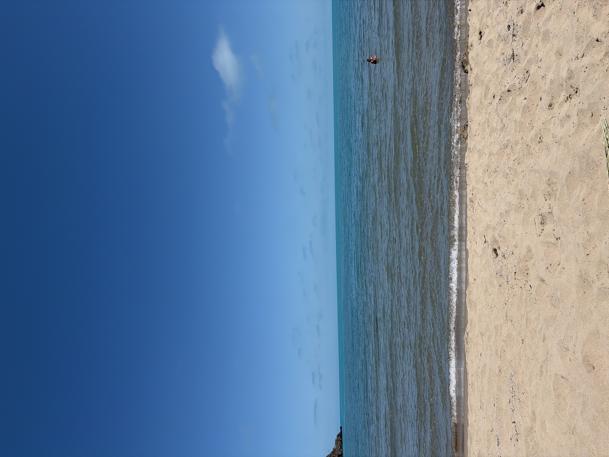 This is a vertical, eye-level, full-shot photograph taken at a beach in Rezovo, Bulgaria. The image depicts a serene beach scene with a clear blue sky dominating the upper half. A lone person is visible wading in the turquoise waters, which are calm with gentle ripples. The foreground is a sandy beach with subtle footprints and debris. A rocky outcrop juts out from the sea on the far left, and a wispy cloud is present in the sky. The overall atmosphere is peaceful and tranquil.