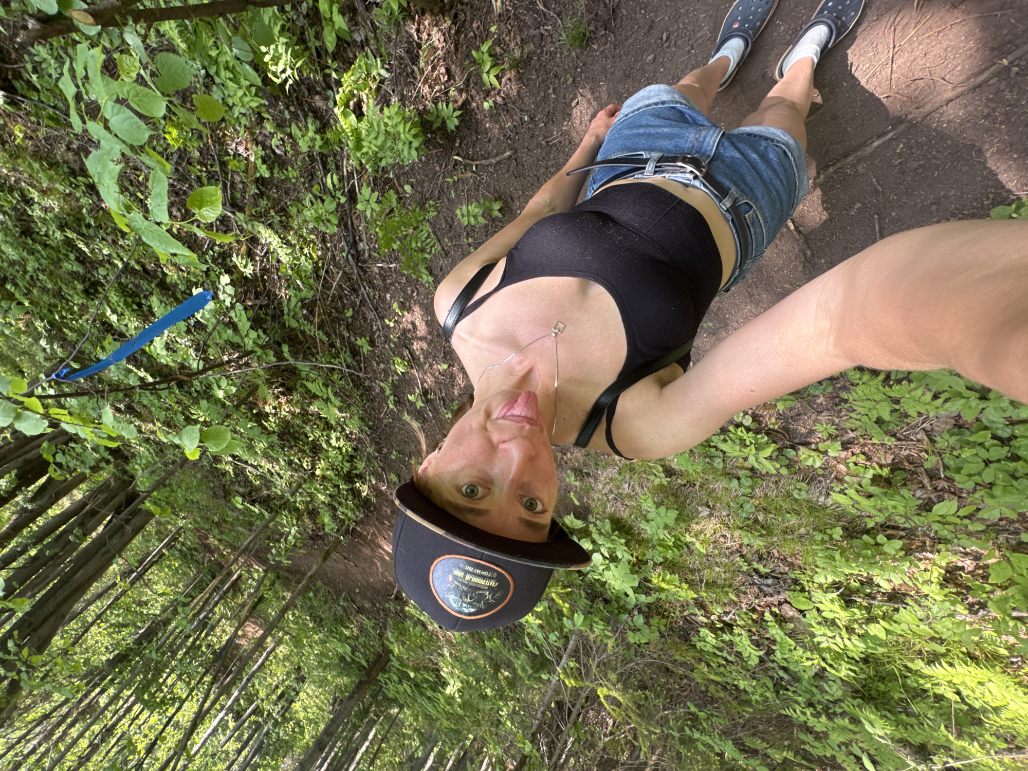 A woman stands on a dirt path in a forest. She wears a black tank top, denim shorts, and Crocs. She has a black baseball cap with a logo, and her tongue is sticking out. A blue trail marker is visible on a tree branch. The location is not specified as the city of Чёрная, Russia.