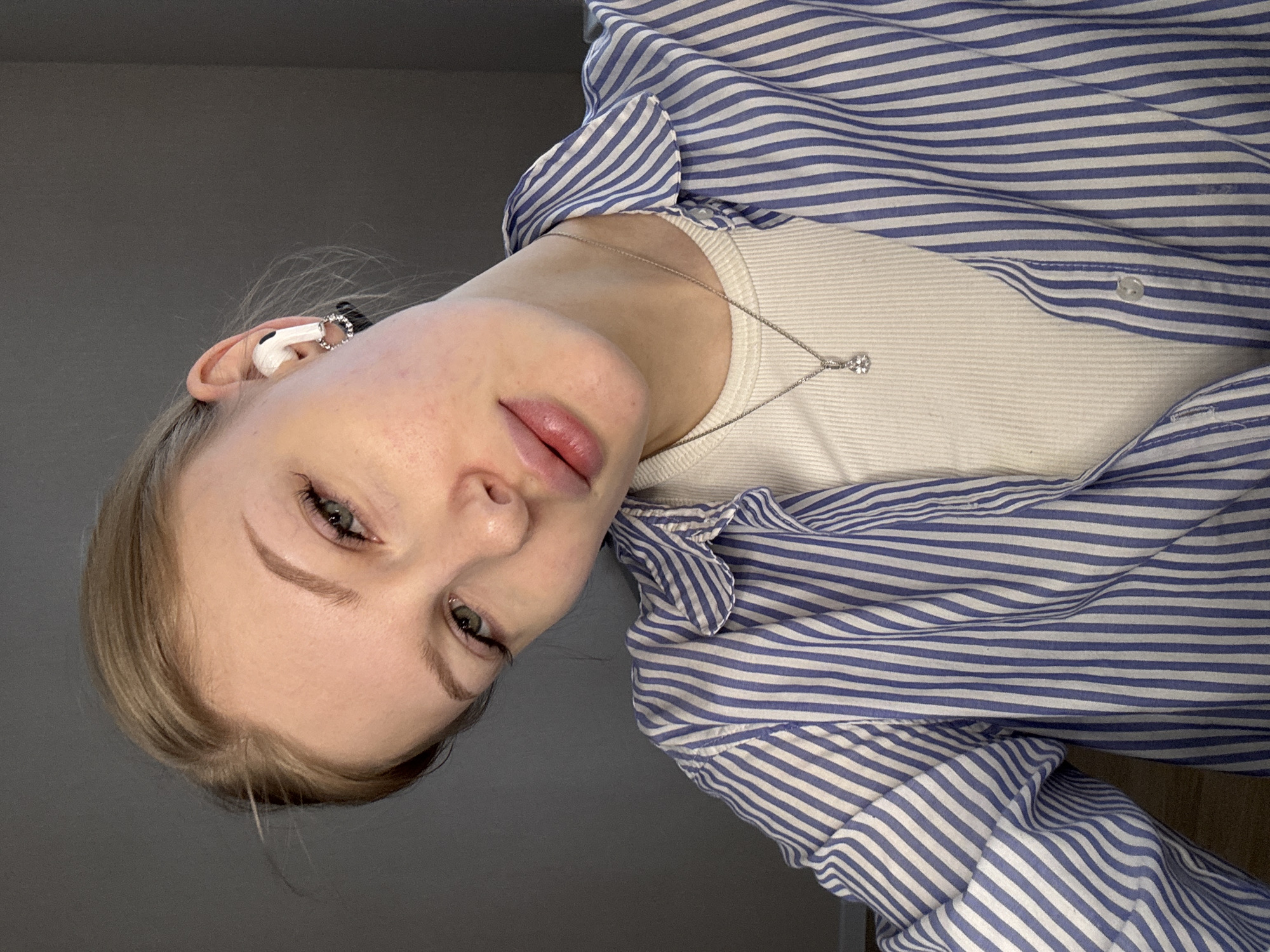 A young Caucasian woman with fair skin and blonde hair is seen in a close-up, slightly angled, indoor shot. She is wearing a white tank top underneath a blue and white striped button-down shirt. A silver necklace with a pendant is visible around her neck. She is wearing an earbud in her left ear. The background is a plain grey wall. There is no indication of location in the city of Perm, Russia.