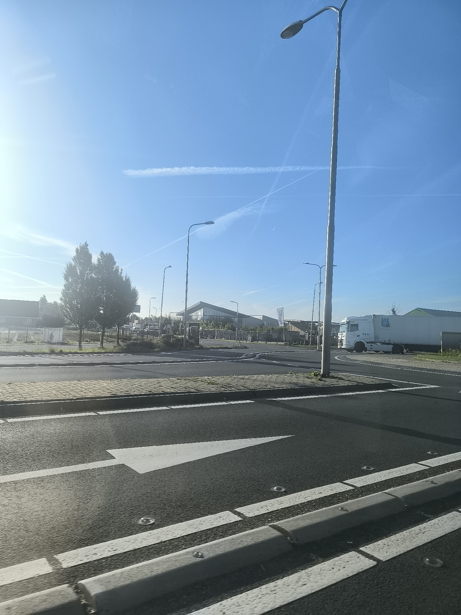 The image shows a bright, sunny day in Boskoop, Netherlands, with a clear blue sky and some wispy clouds. In the foreground, there is a road with a white arrow painted on it, indicating a direction. The road is bordered by a paved walkway and a concrete curb. In the background, there are several buildings, including a modern-looking structure with large glass windows. A white truck with a trailer is parked on the right side of the image. There are also trees and streetlights visible. The overall scene appears to be an industrial or commercial area.