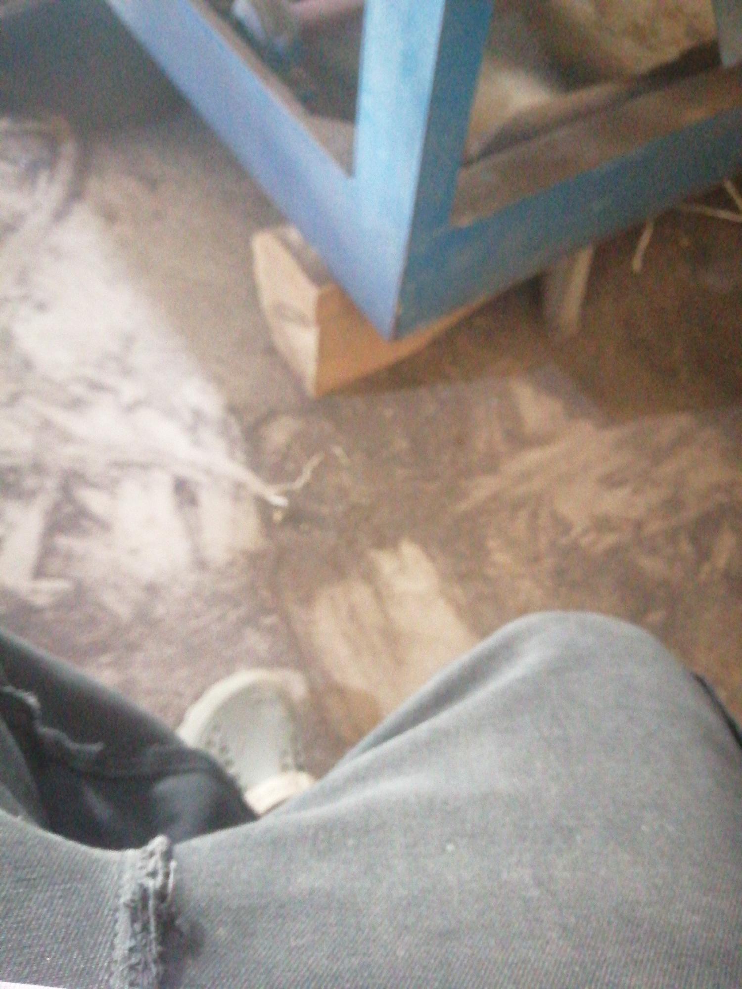 The image shows a close-up view of a person's legs, wearing dark denim jeans and a light-colored shoe. The person appears to be sitting or resting, as their legs are bent. In the background, a blurred blue structure, possibly part of a piece of furniture or equipment, is visible. The environment appears to be indoors with a floor that has a textured surface. The image is unfocused, particularly in the background, making it difficult to discern specific details of the surroundings.