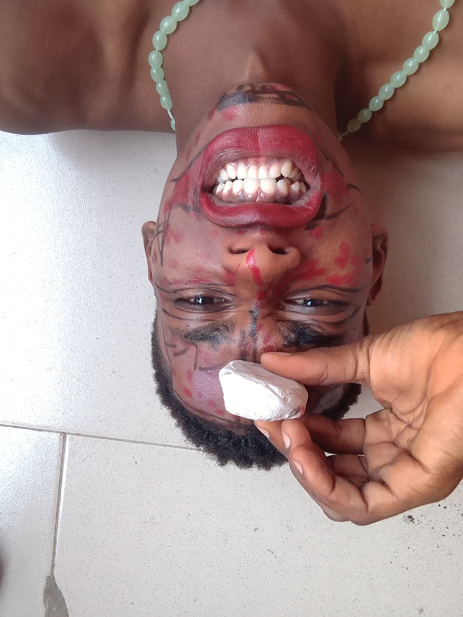 A close-up, high-angle shot depicts a person's inverted head and upper torso, lying on a light-colored, tiled floor. The person, who appears to be male, wears a light green beaded necklace. Their face is covered with red and black markings, resembling tribal paint or theatrical makeup. Bright red lipstick is applied to their widely open mouth, revealing a full set of white teeth in a broad, perhaps intense, smile or grimace. Their eyes are open, looking towards the camera. A hand, with skin tone consistent with the subject, holds a small, white, irregularly shaped object (possibly chalk or a stone) near the person's nose and forehead. The background consists of a simple tiled floor with visible grout lines. The image has a theatrical or ceremonial feel due to the facial adornments and dramatic expression. There is no visible text.