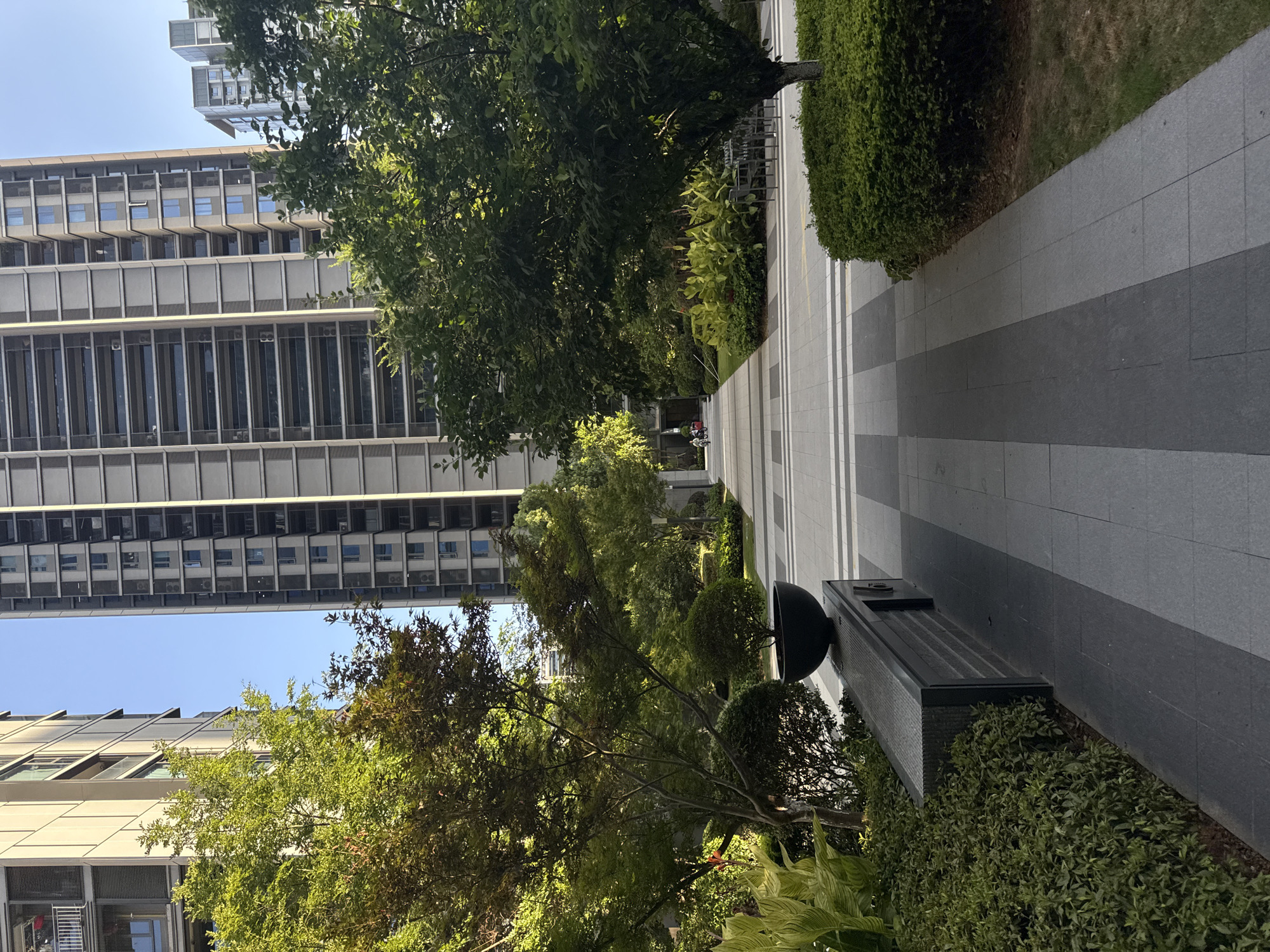 The image depicts an outdoor paved walkway flanked by lush green trees and manicured bushes. Two tall, modern apartment buildings with numerous windows and balconies dominate the background, suggesting an urban residential setting. The walkway is paved with rectangular tiles in alternating shades of gray and white, creating a geometric pattern. A dark gray bench with a stone surface is visible on the left side of the walkway, next to a black planter holding a small, spherical tree. Further down the walkway, two individuals are seen on bicycles, indicating activity in the area. The environment appears to be a well-maintained communal space within a residential complex. This scene is likely located in Yiwu, China, a city known for its extensive residential developments and green spaces.