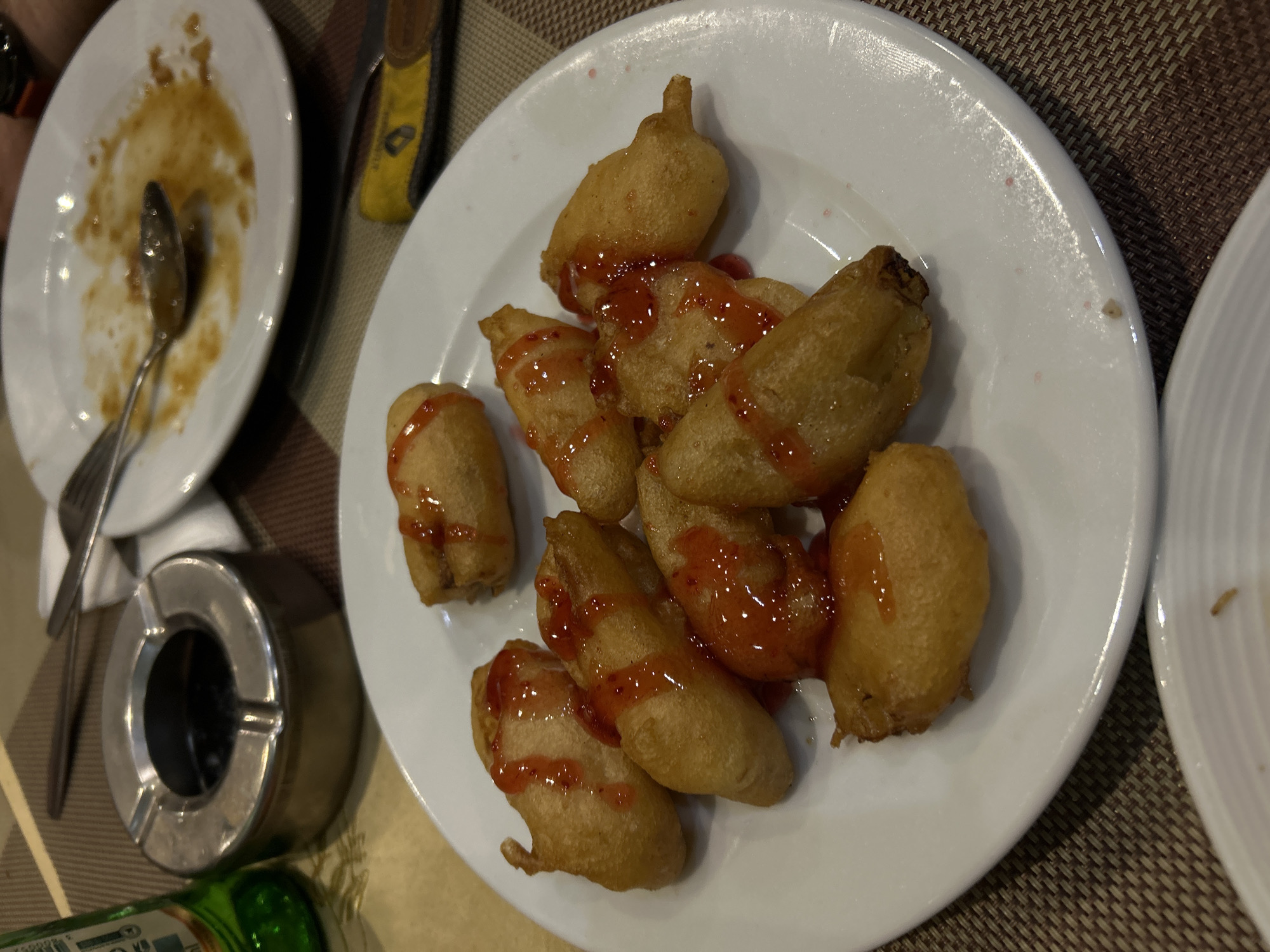 The image shows a plate of fried food items, possibly bananas or similar fruit, drizzled with a red sauce, likely strawberry or raspberry. The plate is white and is on a table with a woven placemat. There is another plate to the left with food residue and a spoon. Also to the left is an ashtray. The lower part of a green bottle is visible in the bottom left corner. The location is possibly a restaurant in Varna, Bulgaria, given the context. No people are visible in the image. The scene depicts a meal setting after some food has already been consumed.