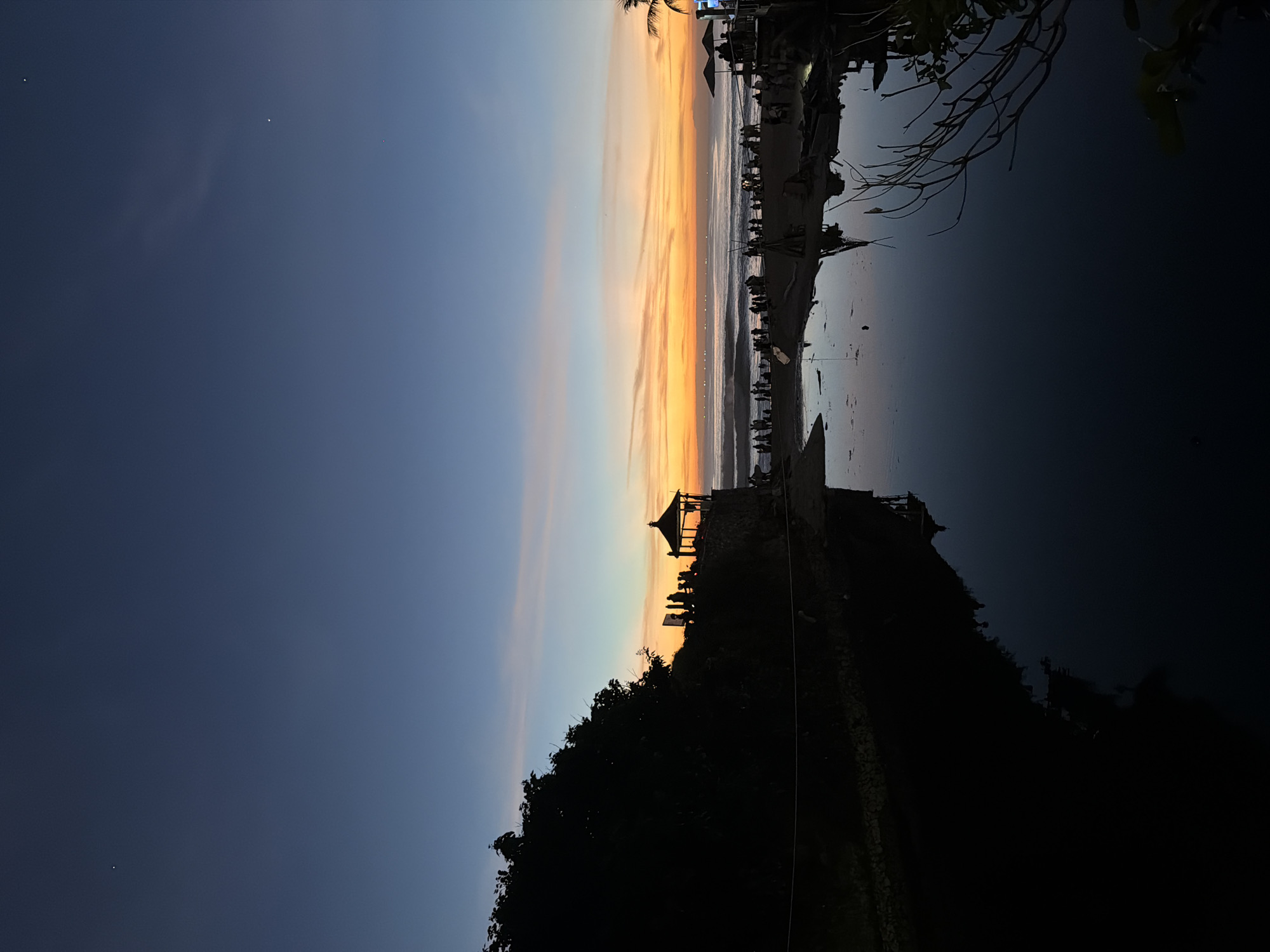 This image shows a sunset scene at the Uluwatu Temple in Kuta Selatan, Indonesia. The sky transitions from blue to orange. A silhouetted structure, presumably a temple section, sits atop a dark, rocky cliff. Several people are visible in silhouette near the temple structure. The ocean reflects the colors of the sunset. A beach area with more people is visible in the distance, with palm trees.