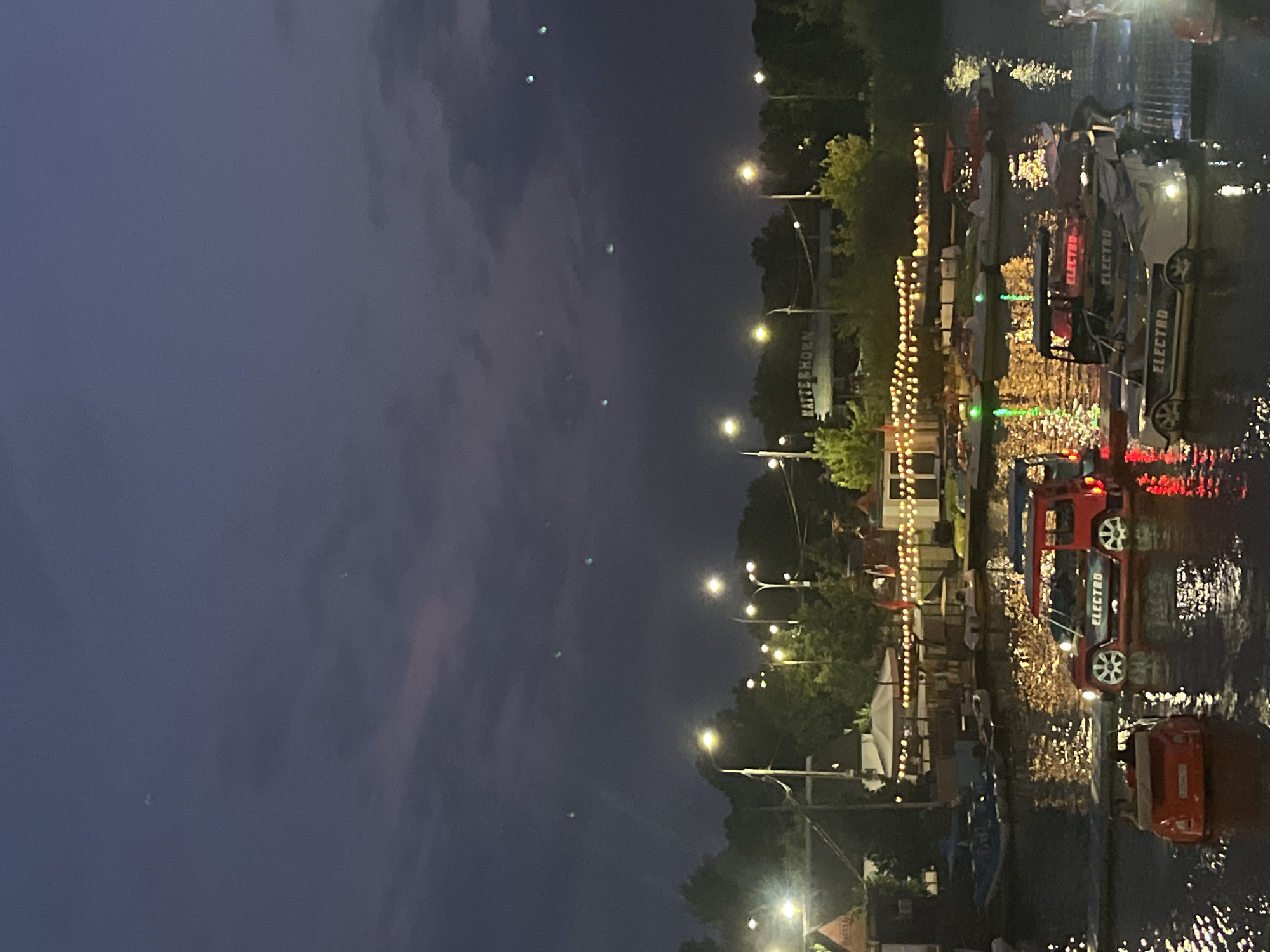 The image captures a dusk scene at an amusement park or recreational area in Krasnodar, Russia, possibly located along the Kuban River. The foreground is dominated by a body of water, likely a pond or a calm section of the river, where several pedal boats designed to resemble cars are floating. Two red pedal boats shaped like cars are prominently featured, one in the lower left corner and another in the center. The boat in the center has "ELECTRO" written on its side. Another pedal boat, white and also designed like a car with "ELECTRO" written on it, is to the right of the red car-shaped boat.
In the background, across the water, there is a strip of land with trees and buildings. A sign with the text "MATTERHORN" is visible on one of the buildings. String lights are illuminated on a structure that appears to be a cafe or seating area, casting a warm glow on the water. Streetlights are also lit, illuminating the pathways and trees. The sky is a deep blue, characteristic of twilight, with some wispy clouds. Several small, bright green lights are scattered across the sky, possibly reflections or atmospheric phenomena. The overall atmosphere suggests a leisurely evening at an entertainment venue. There are no people clearly visible in the image, though the presence of the pedal boats implies recreational activity.