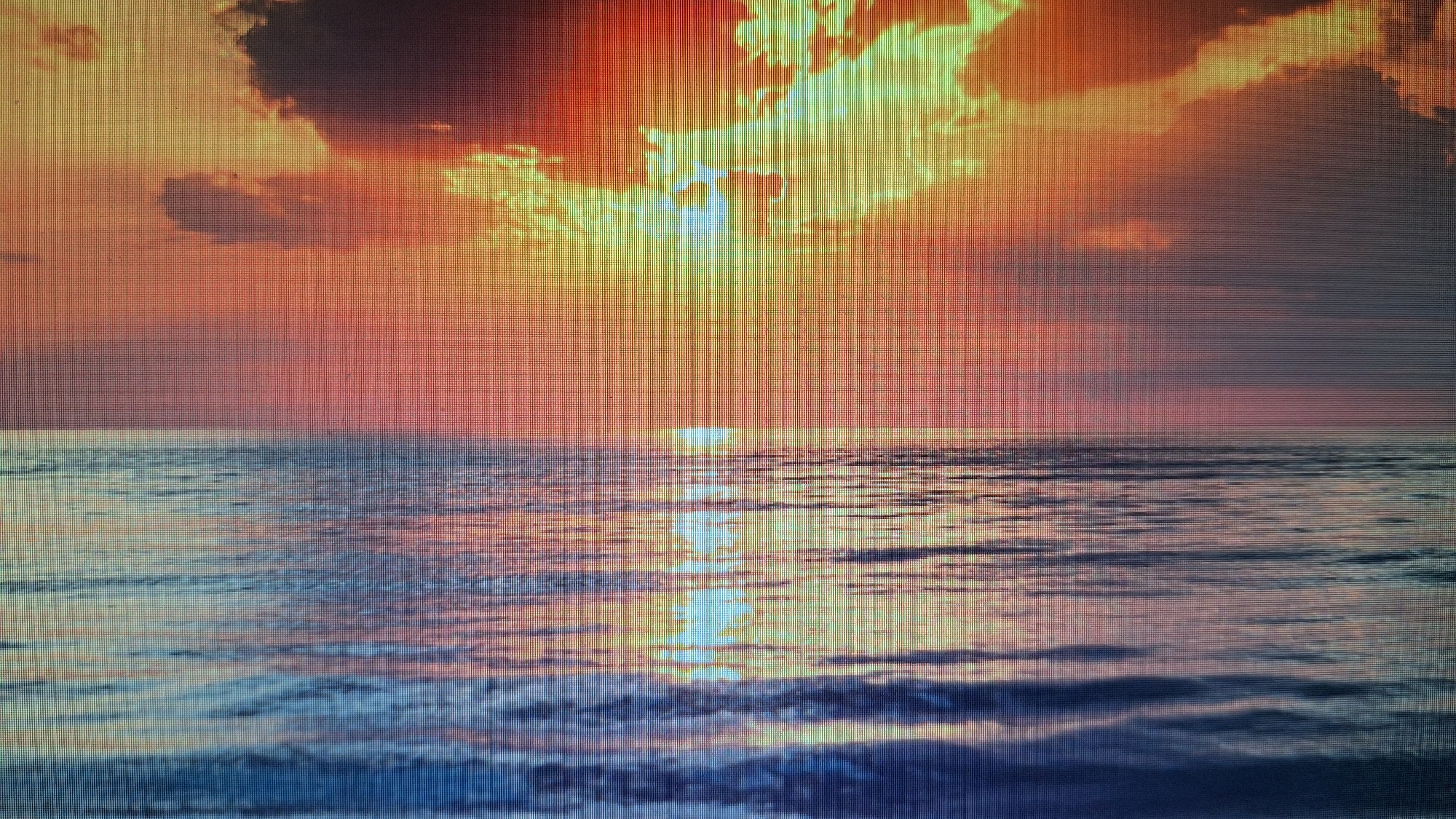 The media file displays a vivid seascape at either sunrise or sunset, characterized by a dramatic and colorful sky reflecting on calm water. The scene is dominated by warm hues of orange, red, and yellow in the sky, with areas of darker clouds interspersed. Bright beams of sunlight pierce through the clouds at the center, creating a striking visual effect.
The horizon line cleanly separates the sky from the ocean. The surface of the water is relatively calm, showing gentle ripples and a prominent, shimmering path of reflected sunlight extending from the horizon towards the viewer. The water itself takes on various shades of blue, purple, and orange due to the reflections from the sky. No people or man-made objects are visible in the scene.
The image itself exhibits a distinct pixel grid pattern across its entirety, which is characteristic of a photograph taken of a digital display screen rather than a direct photograph of the natural scene. This suggests the image is a reproduction of an original digital artwork or photograph shown on a screen. No visible text is present within the image.
While the location context provided is Bankya, Bulgaria, the depicted scene is clearly an expansive seascape, which does not align with Bankya's inland, mountainous geography. Therefore, the image portrays a generic ocean sunset/sunrise, potentially captured on a screen in Bankya.