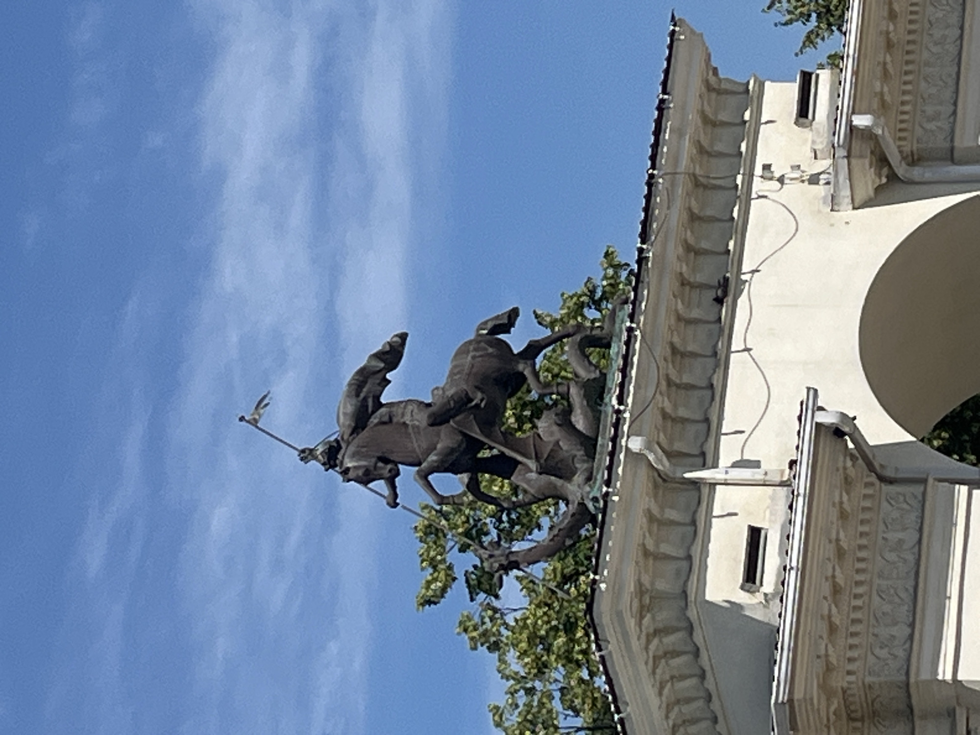 This image depicts a monument of Saint George slaying the dragon, located atop an ornate building structure in Krasnodar, Russia. The sculpture is made of bronze and features Saint George on horseback, piercing the dragon with a lance. The building itself is painted white and has a decorative facade with arched openings. Green trees are visible behind the monument, and the sky is a clear blue with some scattered clouds. The overall scene suggests a bright, sunny day.