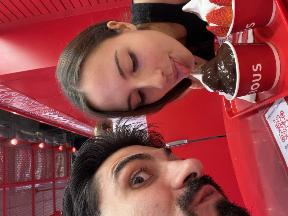A close-up, selfie-style photograph depicts two individuals inside a "Sunnylicious" dessert establishment located in Bangkok, Thailand. On the left, a male subject with dark hair, a beard, and a mustache looks directly at the camera. To his right, a female subject with long brown hair, wearing a dark top, leans forward with her tongue extended, actively licking the peak of a dark brown, possibly chocolate, soft-serve dessert presented in a red cup. Next to this, on a red serving tray, is another red cup containing white soft-serve ice cream topped with sliced fresh strawberries. The immediate foreground shows a red table surface with a white QR code sticker. The background features a striking red interior, with a textured red wall displaying white lettering, partially visible as "SUNNYLICIOUS," and overhead red-framed structures with light fixtures.