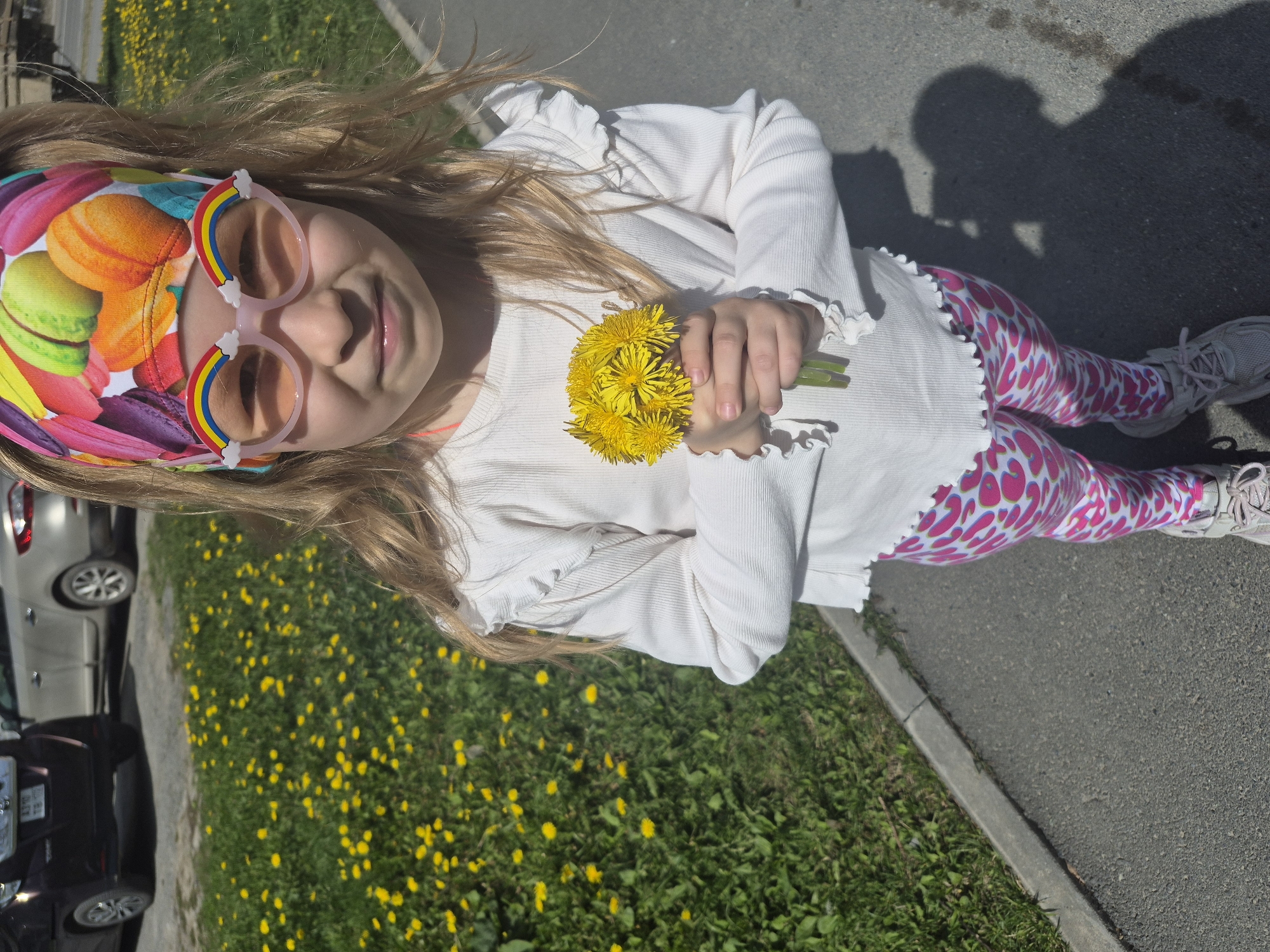 A young girl stands on a sidewalk, holding a bouquet of yellow dandelions. She is wearing a white, long-sleeved shirt with ruffled edges, pink and white patterned leggings, and white sneakers. She has a headband with a colorful macaroon pattern and rainbow-shaped sunglasses. A grassy area with more dandelions is adjacent to the sidewalk. Behind the girl, parked cars are visible. The location is inferred to be Tobolsk, Russia. The girl is smiling.