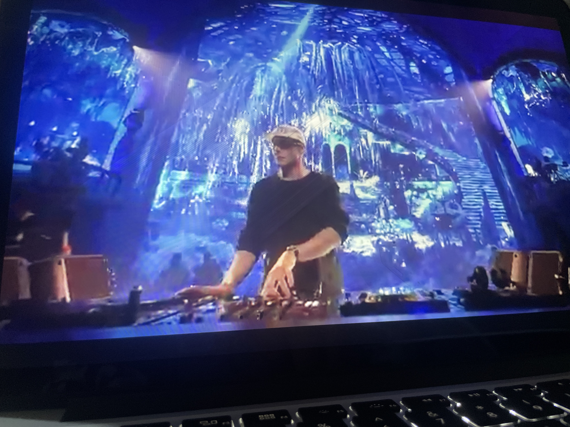 The image shows a person, likely a DJ, at a console in front of a large, illuminated stage backdrop. The backdrop appears to be a stylized representation of ice formations or crystalline structures, illuminated with vibrant blue and white lights, creating an ethereal, possibly futuristic or fantasy-like atmosphere. The DJ is wearing a dark long-sleeved shirt and a light-colored cap and sunglasses, with their hands on the DJ equipment. There are other figures visible in the background, appearing as silhouettes or out of focus, suggesting a crowd or other stage personnel. The setting is on a stage, indicative of a performance or event. There is no identifiable information within the image to suggest a specific location in Kason, Nigeria.