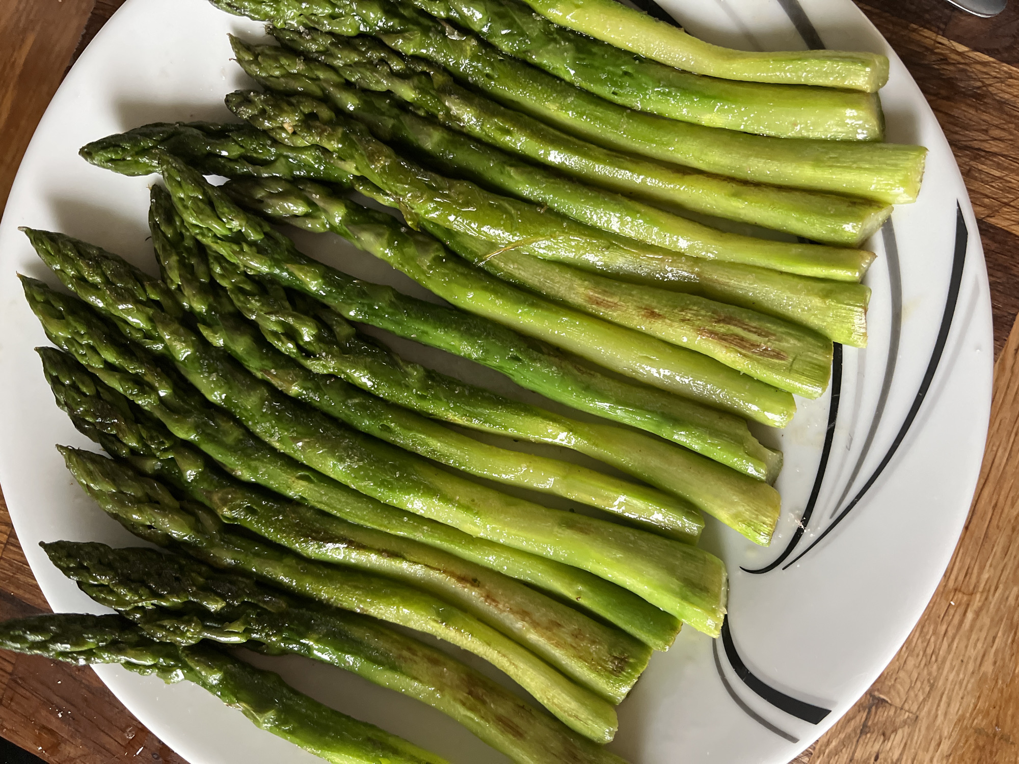 A generous serving of cooked asparagus spears is presented on a white, oblong serving plate. The asparagus is bright green, varying slightly in shade, with some spears appearing lighter towards the cut ends and darker, more vibrant green at the tips. A glossy sheen coats many of the spears, suggesting they are moist from cooking or coated with oil. Close inspection reveals small, sparkling crystals, likely salt, on the surface of some stalks. Occasional light brown or slightly charred marks are visible on the stalks, indicating a cooking method such as roasting or sautéing. The asparagus is arranged in parallel rows, covering most of the plate.
The white plate features a minimalist design with abstract black and gray curved lines or swirls, primarily visible along its right edge. The background consists of a warm-toned wooden surface, possibly a cutting board or table, which is visible in the top right and bottom right corners and appears slightly out of focus. There is no visible text in the image. The scene suggests a prepared dish ready for consumption.