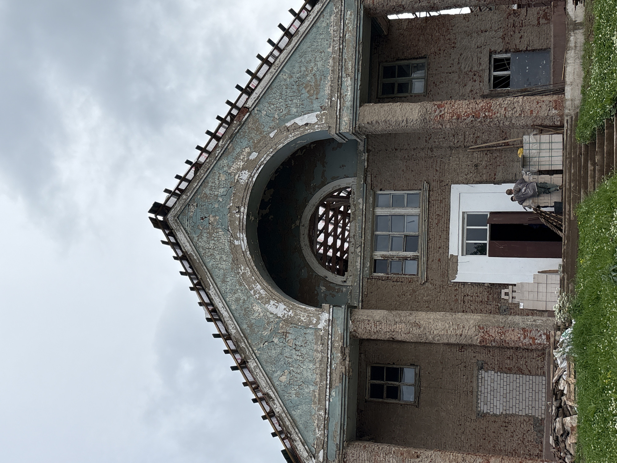 In the city of Kstynino, Russia, an old, dilapidated building is captured under an overcast sky. The structure, possibly a former cultural center or administrative building, exhibits significant decay. The facade is made of brick and stone, with peeling light blue paint adorning a prominent arched section above the entrance. The roof appears to be undergoing repair or has been partially dismantled, with exposed rafters and roofing materials visible. Large, weathered stone columns support the upper architectural elements. Several windows are present, some intact with glass, others broken or boarded up.
At the entrance, a white-framed door stands open, revealing a dark interior. Two individuals, likely construction workers or caretakers, are near the doorway. One person, wearing a dark jacket and light-colored pants, leans against a stack of white building materials, which are wrapped in plastic. The other person, in a lighter jacket and dark pants, stands beside them, also appearing to rest or observe. Several stacks of building materials, including what look like concrete blocks, are arranged on the steps leading up to the entrance. To the left of the entrance, a section of the brick wall has been crudely plastered and partially covered with a new brick pattern. Debris, including wood and stones, is piled on the ground in front of the building, interspersed with patches of green grass dotted with small white wildflowers. The overall impression is one of abandonment and ongoing, possibly rudimentary, restoration efforts.