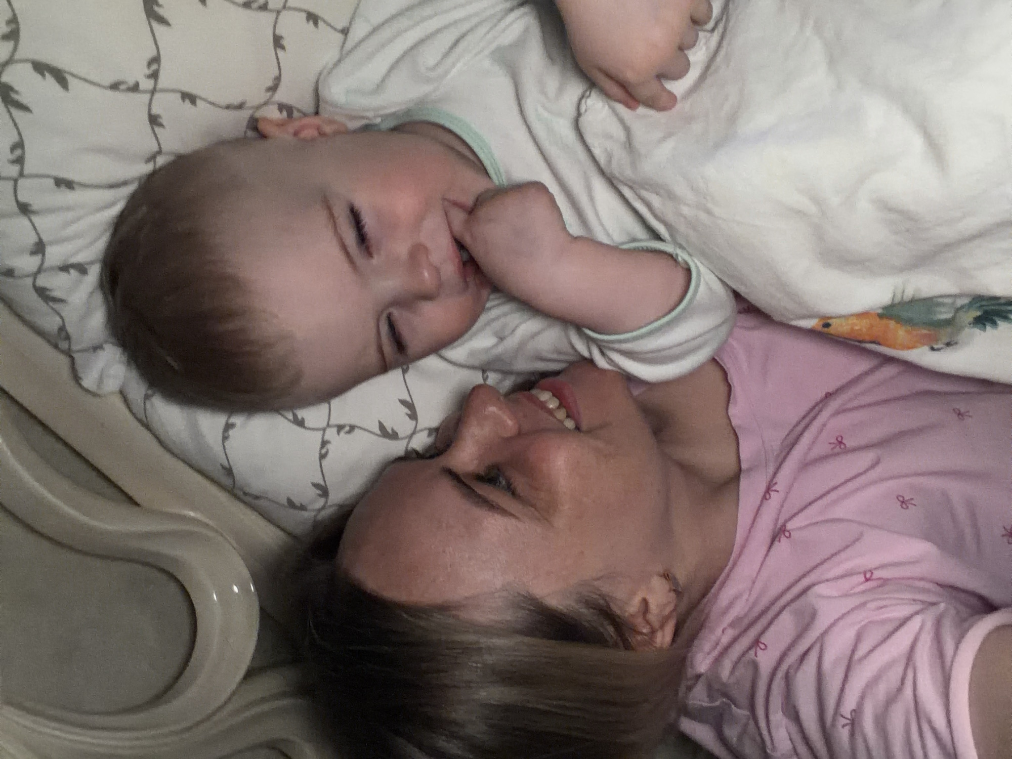 A woman and a baby lie on a patterned bed in an indoor setting. The baby, wearing a white onesie with a light blue trim, has their hand near their mouth. The woman, wearing a pink top, is smiling and looking up at the baby. A portion of a light-colored bed frame is visible on the left. The location is not discernible from the image. The city of Votkinsk, Russia cannot be determined from the image.