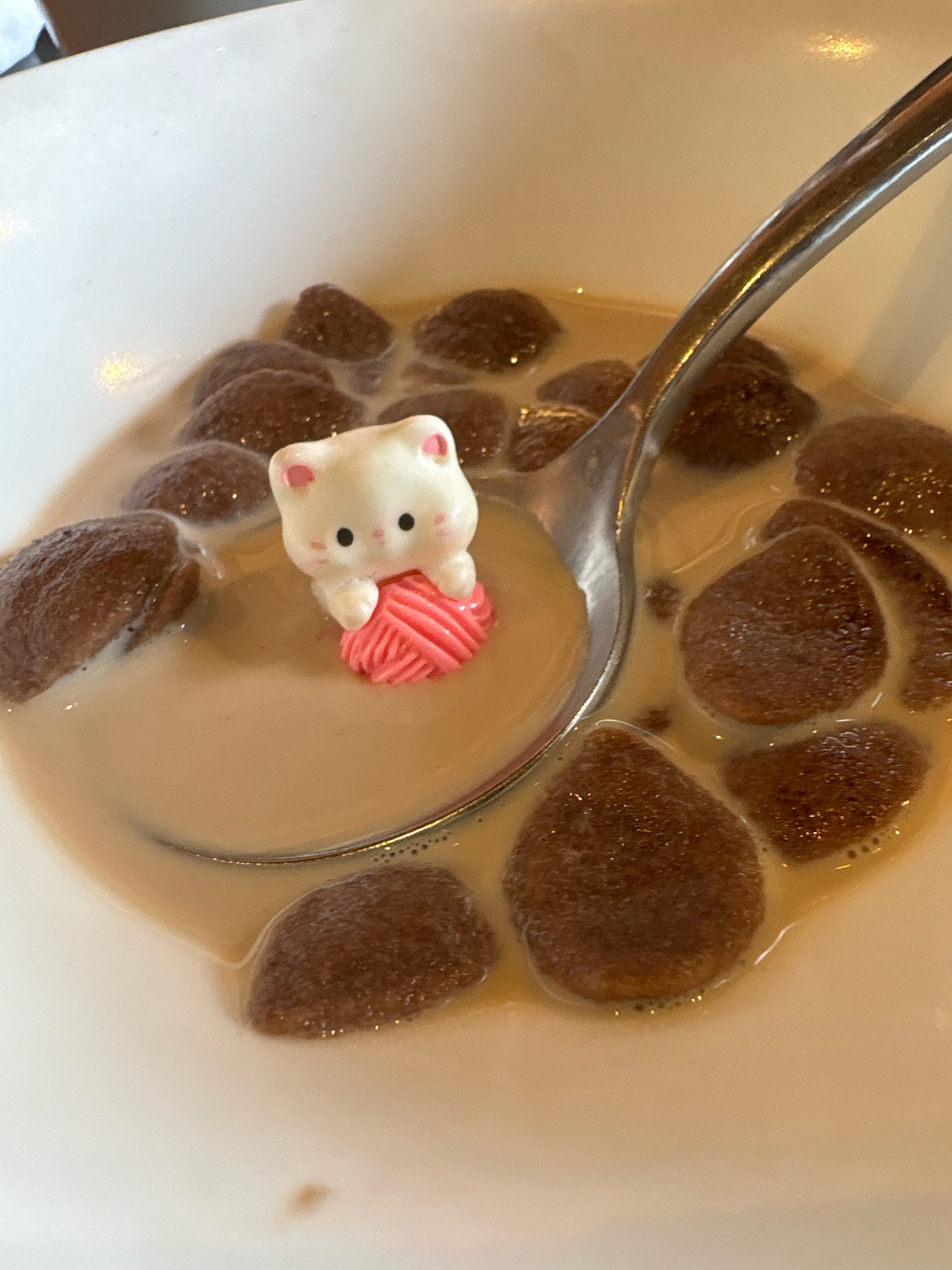 A white ceramic bowl contains a light brown liquid, likely milk or a similar beverage, with several dark brown, rounded objects resembling cereal or small pastries submerged in it. A silver spoon rests in the bowl, partially submerged and positioned to the right of center. Resting on the spoon, within the liquid, is a small, white figurine of a cat with black eyes and pink ears and cheeks. The cat figurine is holding a small, pink, ball of yarn. The background is out of focus, but suggests a bright, possibly sunlit environment. The image is a close-up shot, focusing on the contents of the bowl and the figurine. There is no information in the image to suggest a location in Vagator, India.