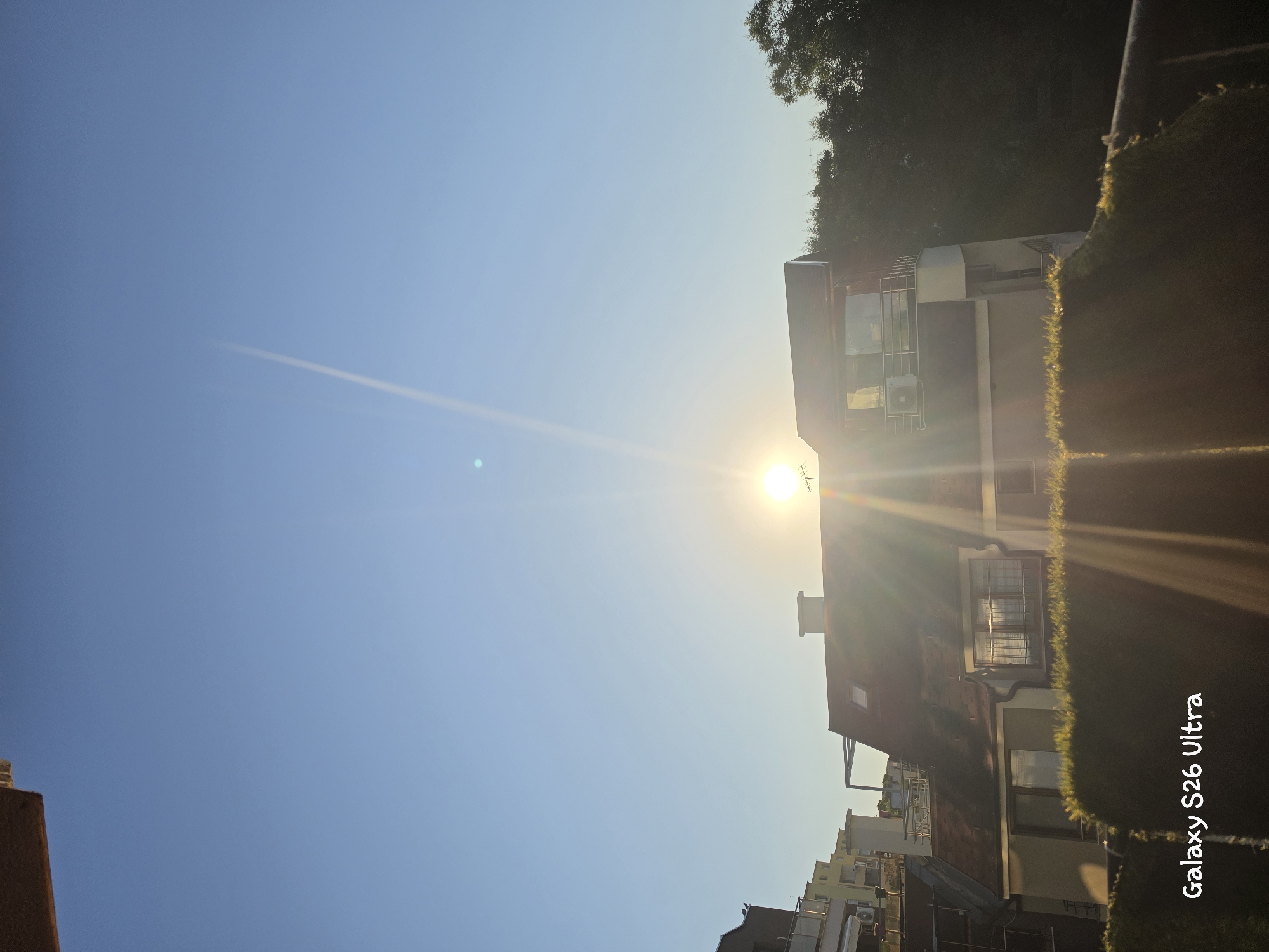 A bright sun is setting over a residential area in Plovdiv, Bulgaria. The sun's rays create a lens flare that stretches across the sky and down towards a building with a red tiled roof. The building has windows, a chimney, and a balcony with railings. In front of the building is a dense green hedge. In the foreground, the text "Galaxy S26 Ultra" is visible, likely a watermark from a smartphone. The sky is clear and blue, with a faint airplane contrail visible high up. The image captures a typical urban scene during golden hour.