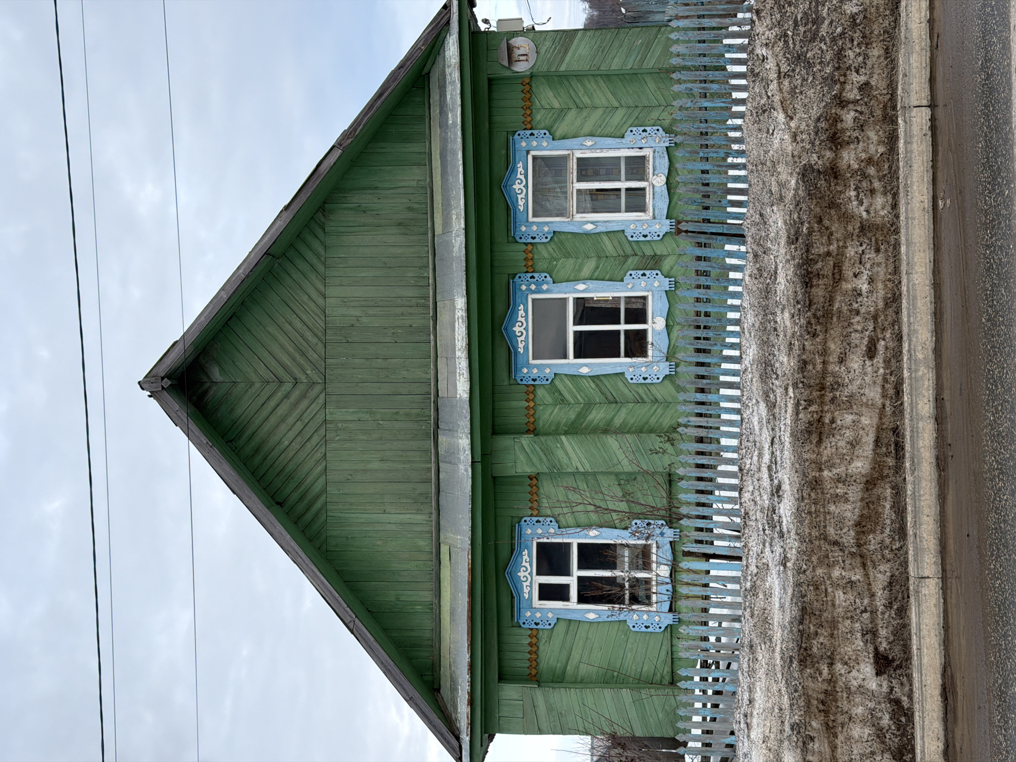 This image displays a two-story, gabled wooden house in Chaikovsky, Russia, viewed from a frontal perspective under an overcast sky. The house exterior is painted faded green, featuring horizontal wooden siding on the lower story and diagonal wooden planks on the gable. Three evenly spaced white-framed windows with multiple panes are present on the facade, each embellished with ornate, carved light blue and white wooden nalichniki (window surrounds). A light brown decorative band runs horizontally beneath the eaves and continues between the windows. A light-colored utility box and a small, rectangular birdhouse-like structure are affixed to the upper right section of the house wall. A weathered, light blue wooden picket fence stands in front of the house. Immediately foregrounded is a large, dirty snowbank positioned between the fence and the road, indicating a state of melting. A concrete curb separates the snowbank from a wet, dark asphalt road that occupies the bottom of the frame. Sparse, bare tree branches are visible behind the fence and in front of the house. Two dark utility wires traverse horizontally across the grey, cloudy sky in the upper portion of the image. No individuals or discernible actions are depicted.