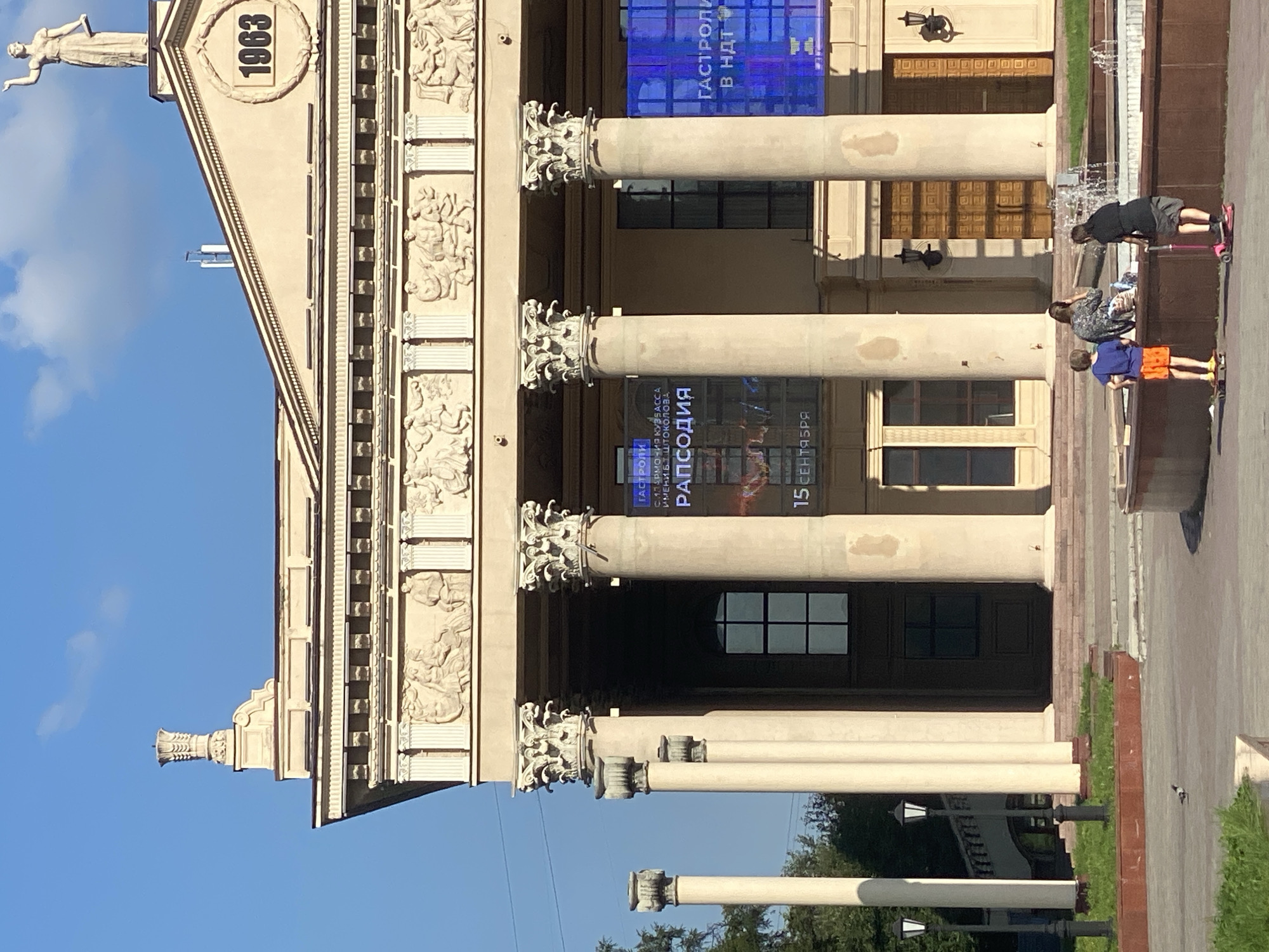 The image shows the facade of a building, likely a theater or cultural center, with classical architectural elements. Large fluted columns with ornate capitals support an entablature decorated with bas-relief sculptures. Above the entablature, the building features a triangular pediment with a decorative urn on the left side and a circular emblem containing the year "1963" on the right. A statue of a standing woman holding an object is positioned at the peak of the pediment.
In front of the building, there are several large banners displayed. One banner, predominantly blue, advertises "GASTRONY" and mentions "V NDT". Another banner, also with blue elements, displays the word "РАПСОДИЯ" (Rhapsody) and the date "15 СЕНТЯБРЯ" (September 15th).
In the foreground, a fountain is visible. Three individuals are present near the fountain: two children on skateboards and an adult seated. One child, wearing an orange shirt and shorts, stands on a skateboard. Another child, wearing a black shirt and shorts, stands on a pink scooter. An adult, dressed in a patterned top, is seated, facing away from the camera, with their hand near their face.
The environment appears to be an urban public space, with trees and streetlights visible in the background. The overall scene is illuminated by bright sunlight, casting shadows. This building is identified as the Novokuznetsk Drama Theatre, located in Novokuznetsk, Russia.