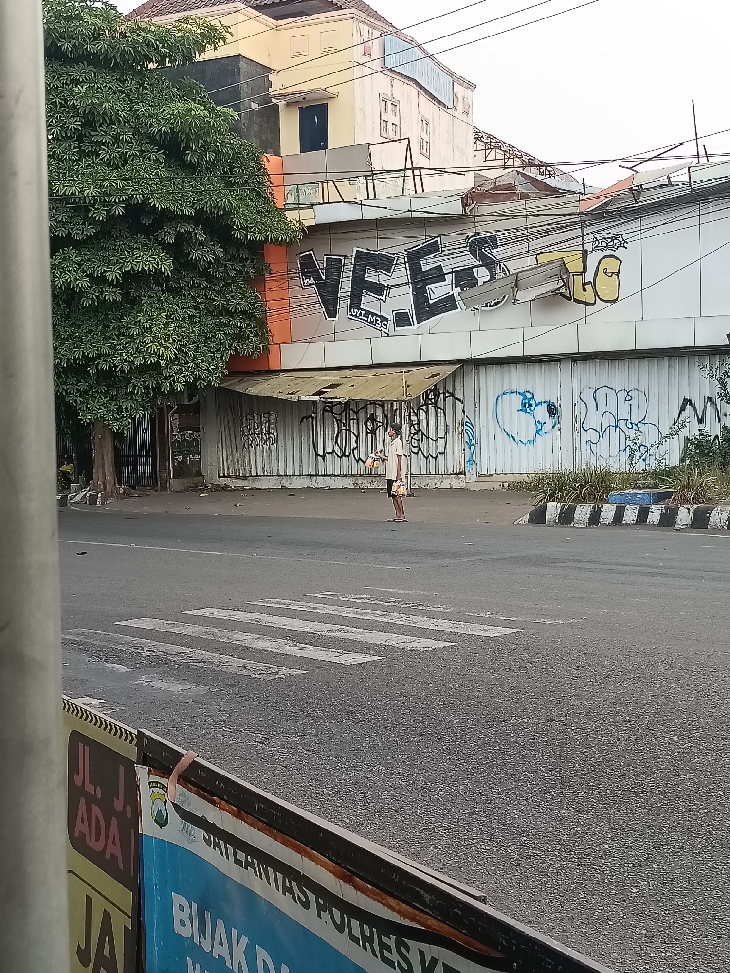 A person stands on a street corner in Kediri City, Indonesia, holding two bags. The person is wearing a light-colored t-shirt and shorts. They are standing in front of a building with graffiti on it. The building appears to be under construction or renovation, with some of its facade damaged. A crosswalk is visible on the street in front of the building. The sky is overcast, and there are power lines visible in the background. The overall scene suggests an urban environment in a state of disrepair.