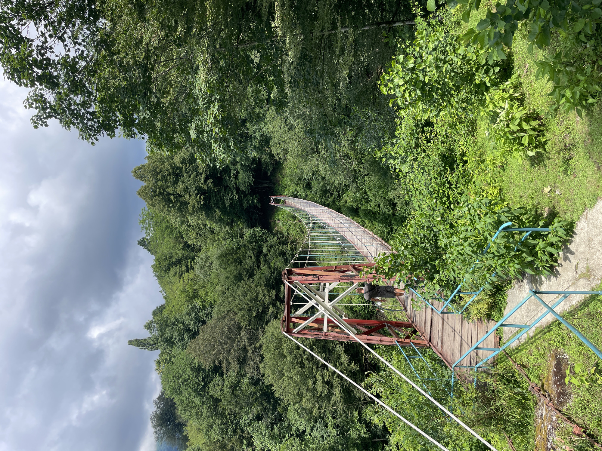 A single person walks away from the camera onto a red and white suspension bridge. The bridge spans a gorge or valley, with lush green trees on either side. The sky is cloudy, with patches of blue visible. The bridge has wooden planks for its walkway, supported by metal cables. The person is wearing a dark t-shirt and shorts. The surroundings are heavily forested, with dense vegetation. The location is identified as Tqwarchal, Abkhazia.