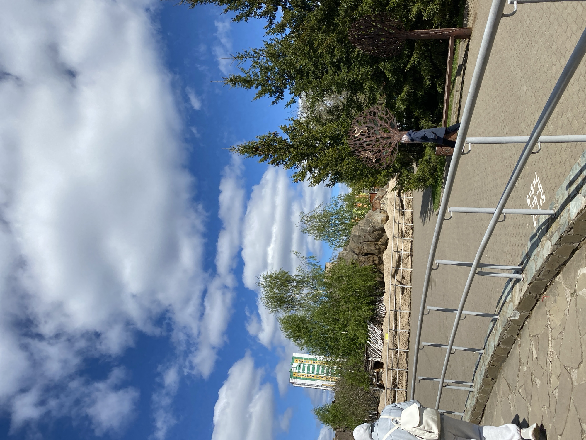 The image depicts an outdoor scene in Izhevsk, Russia. A bright blue sky with scattered white clouds forms the backdrop. In the foreground, there is a paved pathway bordered by a metal railing. Behind the railing, a landscaped area features trees, rocks, and a building in the distance. A person wearing a dark jacket stands beside a decorative metal tree sculpture. The scene appears to be a park or recreational area within the city.