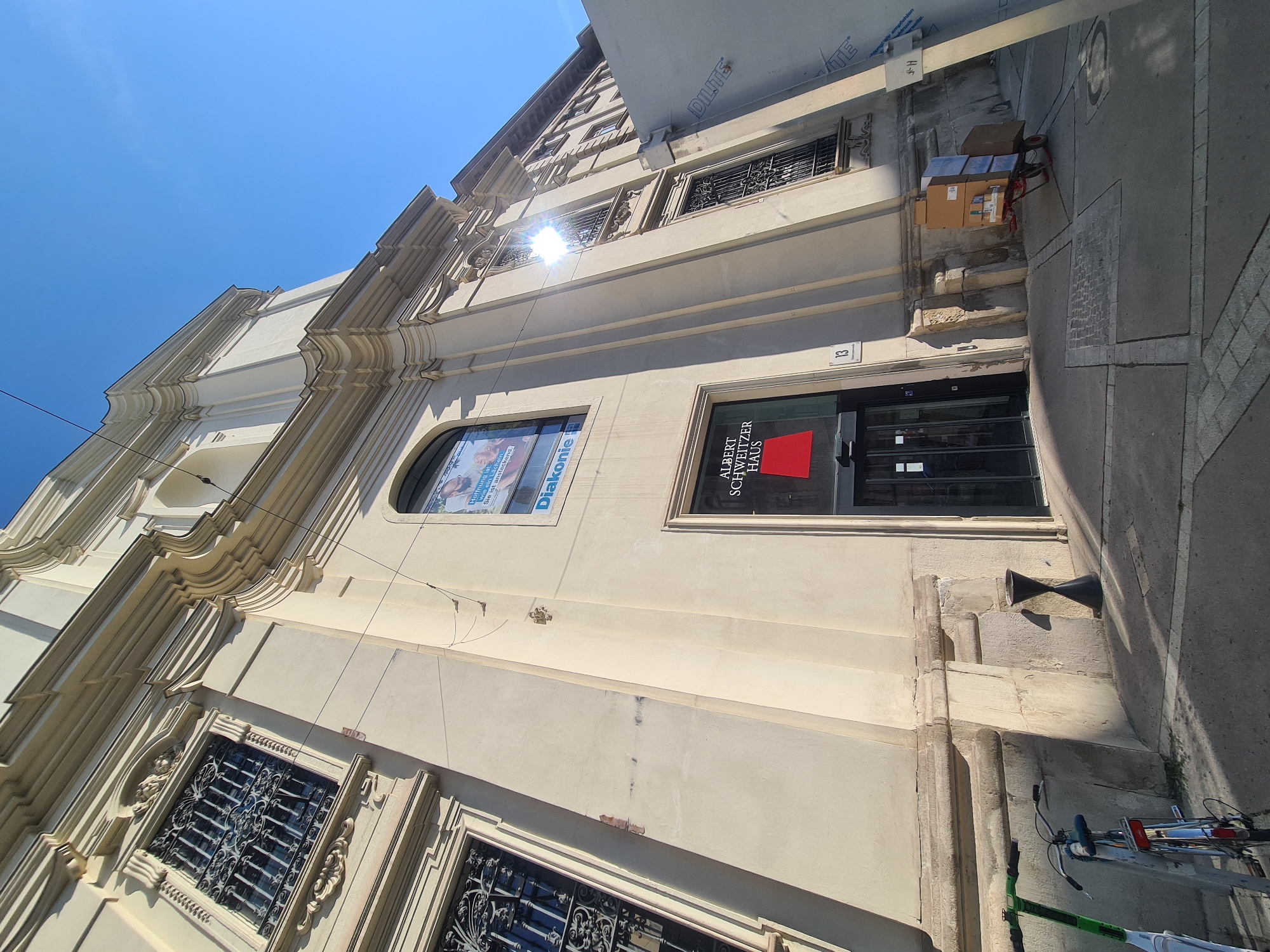 The image depicts the Albert Schweitzer Haus, located at Schwarzspanierstraße 13 in Vienna, Austria. The building is a cream-colored, ornate structure with tall windows featuring intricate ironwork. In front of the building, a glass-doored entrance is visible, with a red sign displaying "ALBERT SCHWEITZER HAUS". To the left of the entrance, there is a window with a poster for "Diakonie". Outside the building, a green electric scooter and a bicycle are parked near the sidewalk. A small black hourglass-shaped bin is also present. Further to the right, a handcart laden with cardboard boxes is positioned on the sidewalk. The sky is a clear, bright blue. The sun is shining, casting strong shadows and creating a glare on the building.