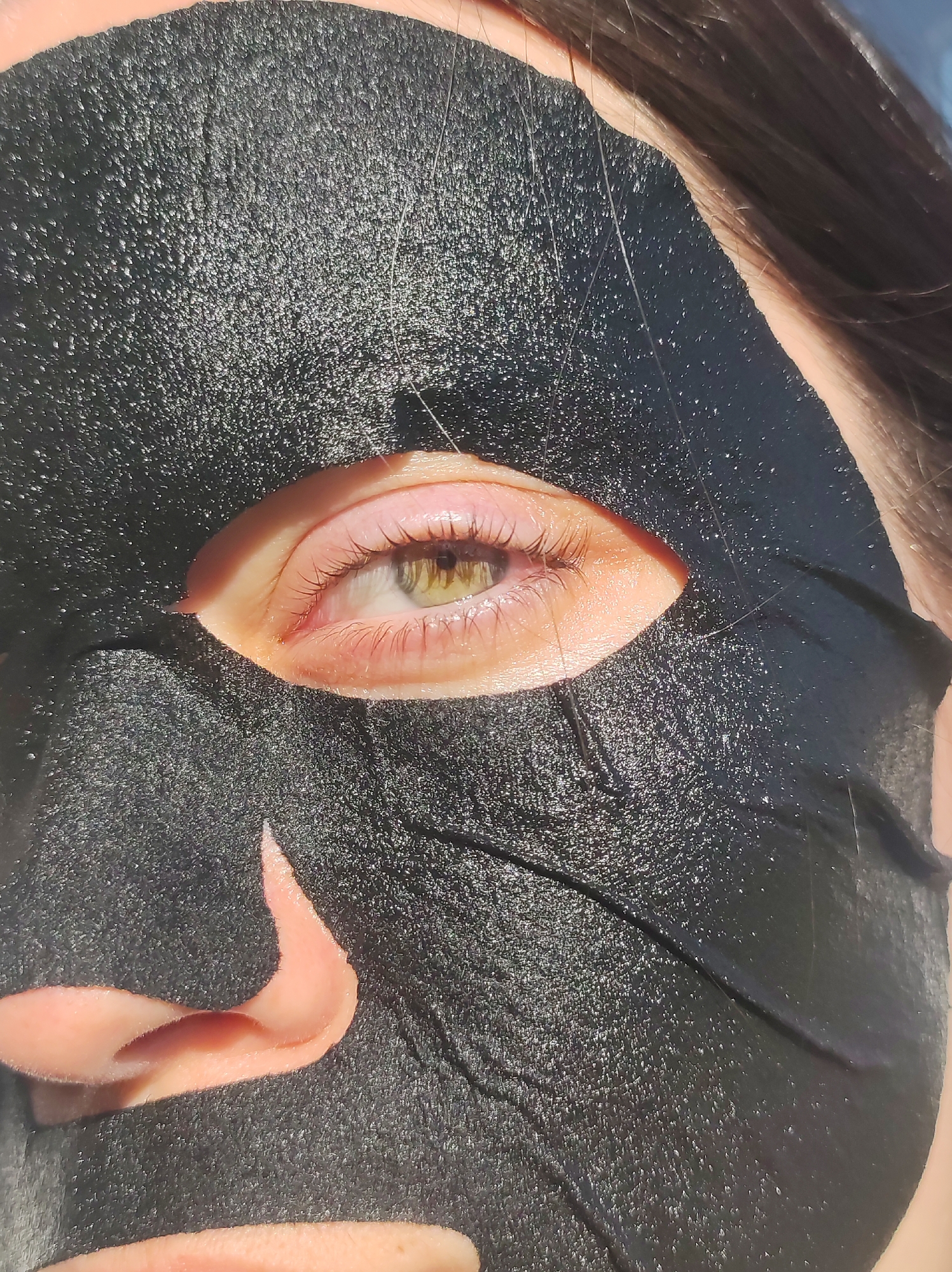 A close-up shot depicts a person's face covered with a black facial mask. The mask is applied to the majority of the face, with only one eye and part of the nose visible. The visible eye is light-colored with eyelashes, and the nose is fair-skinned. The mask appears to be textured, and strands of hair are visible near the edge of the mask. The image is brightly lit, highlighting the details of the mask and the exposed skin. No specific location in Фоки, Russia, can be determined from the image, and no information is available to connect the image to that location.