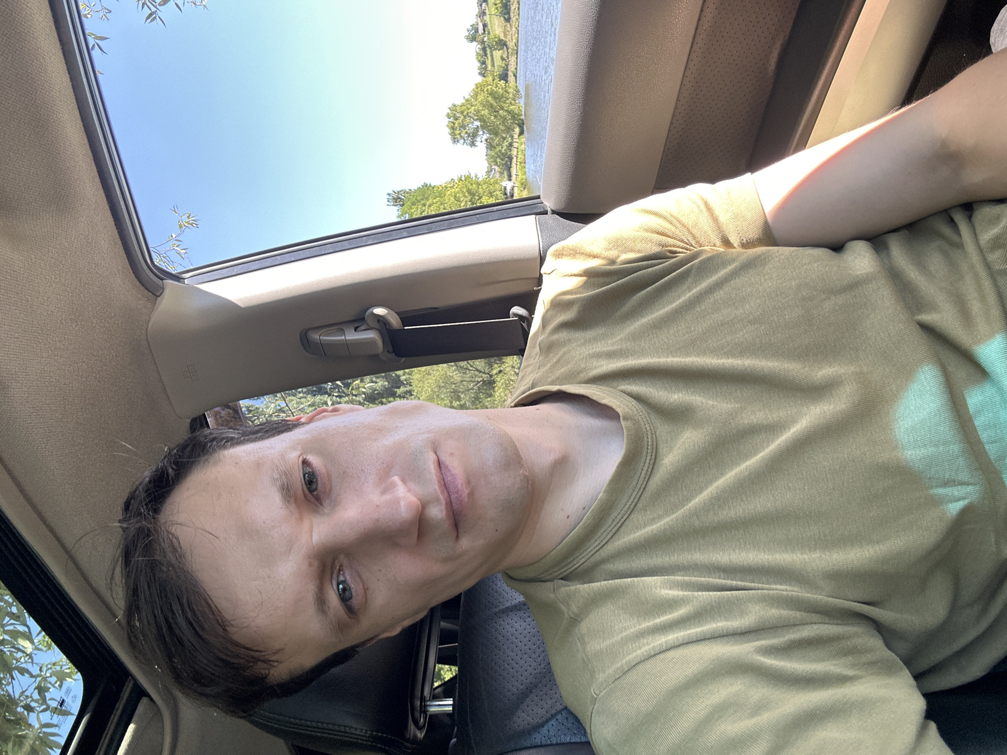 A man is taking a selfie inside a car. The man is wearing a light brown t-shirt. He is looking directly at the camera with a neutral expression. The car has a sunroof open, and through it, we can see the sky and some trees. The side window of the car shows a body of water with trees and buildings in the background. The location is specified as Makiv, Ukraine. The lighting suggests it is daytime, likely late morning or early afternoon.