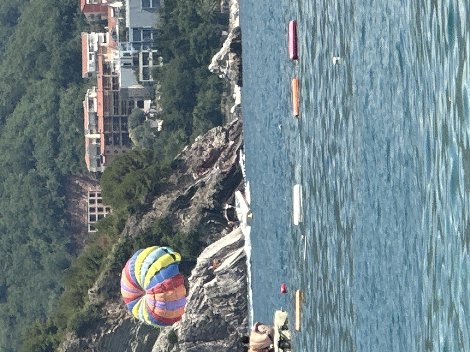 In Boreti, Montenegro, a vibrant parasail, colored with stripes of red, yellow, green, blue, and purple, floats in the air over the azure sea. The parasail is attached to a boat that is in motion, creating a white wake in the water. The scene is set against a backdrop of lush green hillsides dotted with buildings. Rocky outcrops rise from the sea, and on one of these rocks, a person appears to be observing the parasail. In the foreground, several inflatable buoys, one orange and one pink, float on the surface of the water.