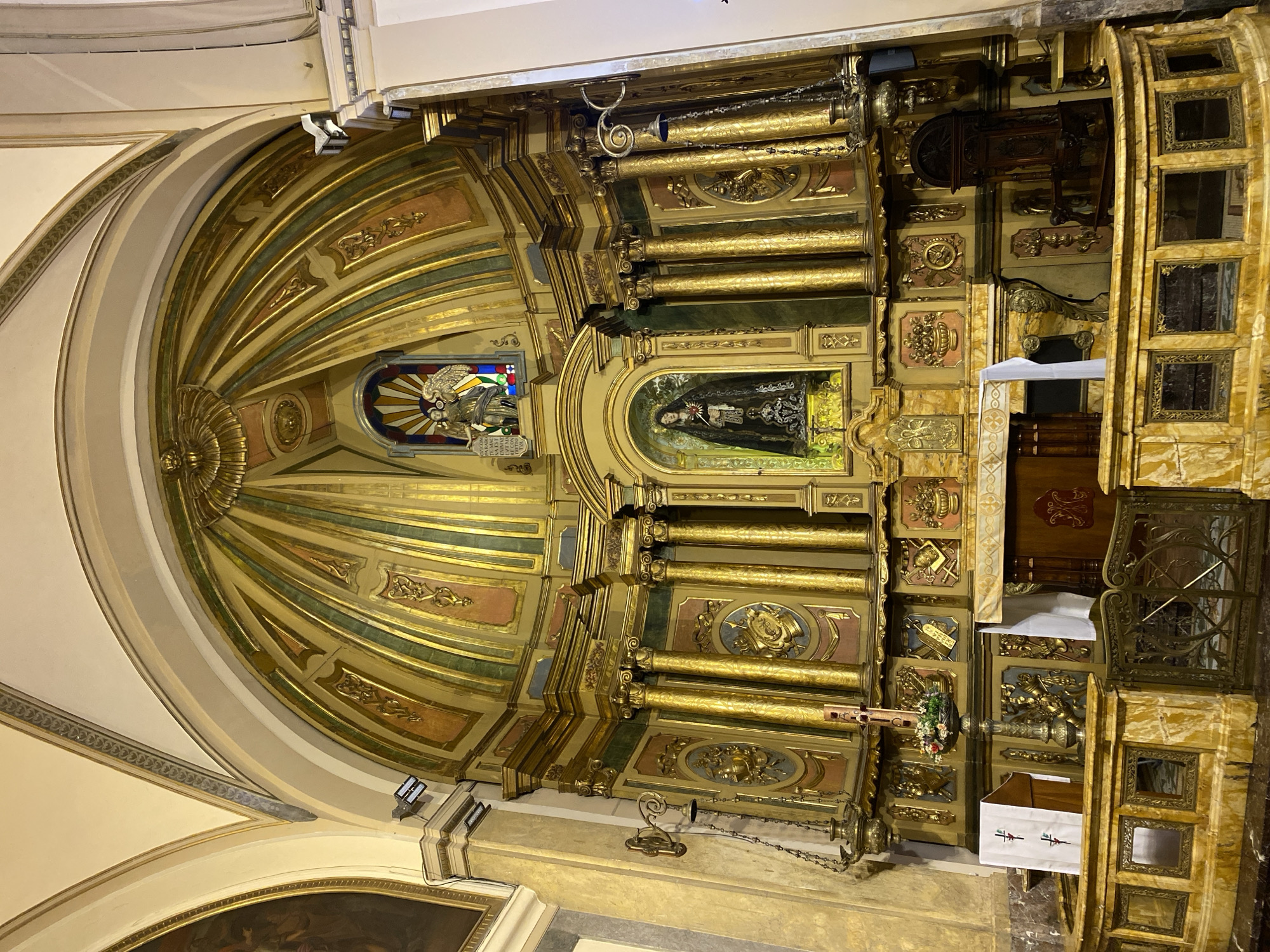 The original image, rotated 90 degrees counter-clockwise, displays the opulent interior of a church or chapel, focusing on the main altar and apse.
The central feature is a highly ornate, multi-tiered gilded reredos. A prominent niche at its center houses a dark-robed statue, possibly the Virgin of Sorrows (Mater Dolorosa), with a heart depicted on her chest. Above and to the left of this central figure, a smaller arch contains a winged statue, likely an angel, positioned against a vibrant stained-glass panel. The reredos is adorned with numerous gilded columns, intricate carvings, and decorative panels in gold, dark green, and reddish-brown.
Above the altar, a grand half-dome or apse ceiling radiates outwards with gilded ribs and painted segments. Modern spotlight fixtures are installed to illuminate the detailed artistry. At the base, what appears to be an altar table or credenza is draped with a white cloth. To the right, a multi-paneled structure with mirrored or glass inserts is visible, along with a decorative bronze hanging ornament resembling a small bell or censer.
A section of stained glass beneath the angel statue displays Latin text, including "GRATIA PLENA" and fragments of "DOMINUS TECUM", likely part of the prayer "Ave Maria, Gratia Plena, Dominus Tecum" (Hail Mary, full of grace, the Lord is with you).
The overall aesthetic is one of rich Baroque or Rococo grandeur, conveying a sense of reverence characteristic of religious architecture in Buenos Aires, Argentina. No specific activity or event is taking place; the image captures the static beauty of the architectural details under artificial lighting.