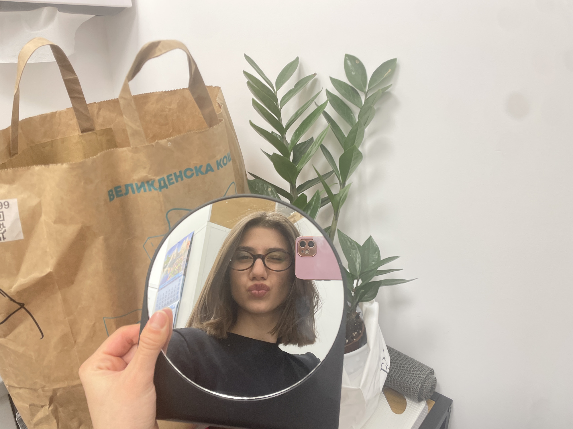 A woman holds a round mirror in her hand, reflecting her face with glasses and a pouting expression. A pink phone case is visible in the mirror. Behind the mirror, there's a brown paper bag with the text "ВЕЛИКДЕНСКА КО" (likely a shortened form of "Великденска кошница," meaning "Easter basket" in Bulgarian), suggesting it's a shopping bag from Bulgaria. The bag is placed near a ZZ plant in a white pot. The background consists of a plain white wall. The scene appears to be indoors. The location is likely Sofia, Bulgaria, based on the Bulgarian text on the bag.