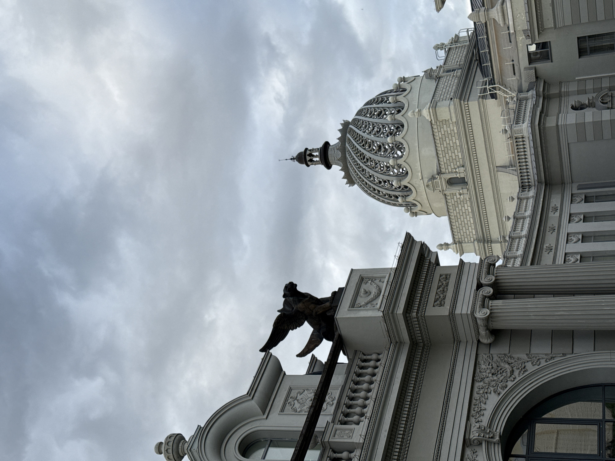 The image shows the National Library of the Republic of Tatarstan in Kazan, Russia, under a cloudy sky. The library features a prominent dome with intricate metalwork and decorative elements. In the foreground, a winged lion sculpture is perched on the edge of a building. The architecture is classical, with columns and ornate detailing. The scene is static and depicts a daytime view of the building's exterior.