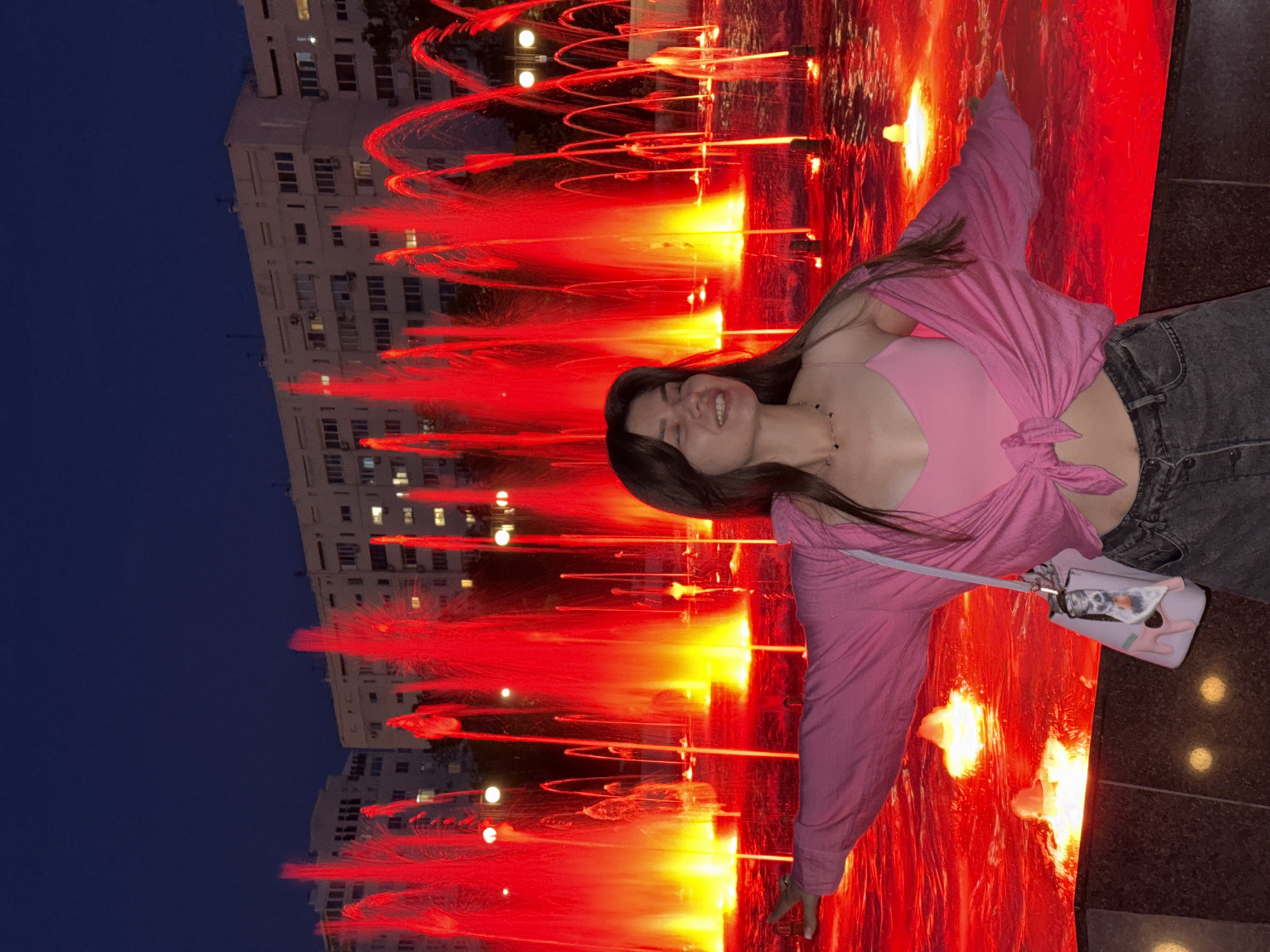 A young woman with long brown hair and closed eyes smiles as she stands with her arms outstretched in front of a vibrant red fountain at night. She wears a pink tank top, a pink open shirt tied at the waist, and dark denim jeans. A light pink purse with a cartoonish charm hangs from her shoulder. The fountain's red lights illuminate the water, creating a dramatic backdrop of cascading sprays. Behind the fountain, a multi-story building with numerous windows is visible against the dark blue night sky. The scene is set in an urban environment, likely a public square or park.
