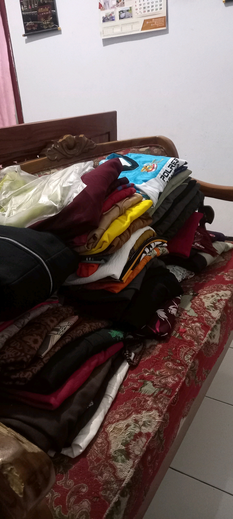In Pontang, Indonesia, a pile of neatly folded clothes rests on a wooden bed frame covered with a red and gold patterned cloth. The clothes, predominantly in shades of blue, red, yellow, and white, are stacked in a disorganized yet compact manner. Some items appear to be shirts, including a blue t-shirt with the word "POLRES" printed on it. A black zippered bag is positioned to the left of the clothing pile. In the background, a white wall is adorned with two framed pictures, one depicting what appears to be a jewelry display and the other a calendar featuring several small photographs. The lighting is dim, casting shadows on the objects.