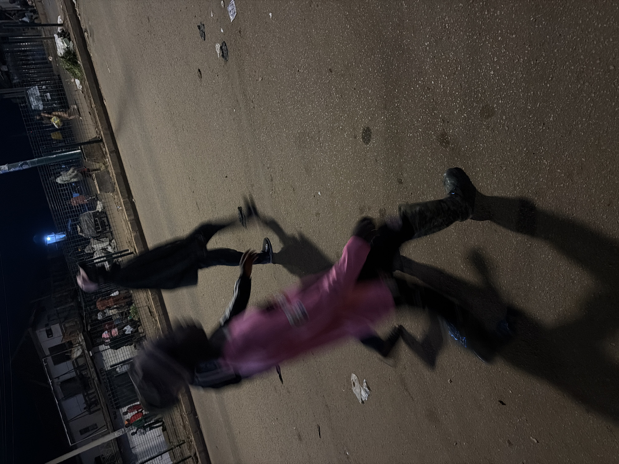 The image depicts a street scene at night in Bukuru, Nigeria. In the foreground, a person is blurred due to motion, wearing a pink top and dark boots, casting a long shadow on the asphalt road. Further back, another person is visible, also blurred and wearing dark clothing. The background shows a row of buildings and a fence. Several other individuals are present in the background, some near the fence and others further back. The road surface appears worn and has some debris scattered across it. The lighting suggests an urban environment at night, possibly illuminated by streetlights or lights from the buildings.