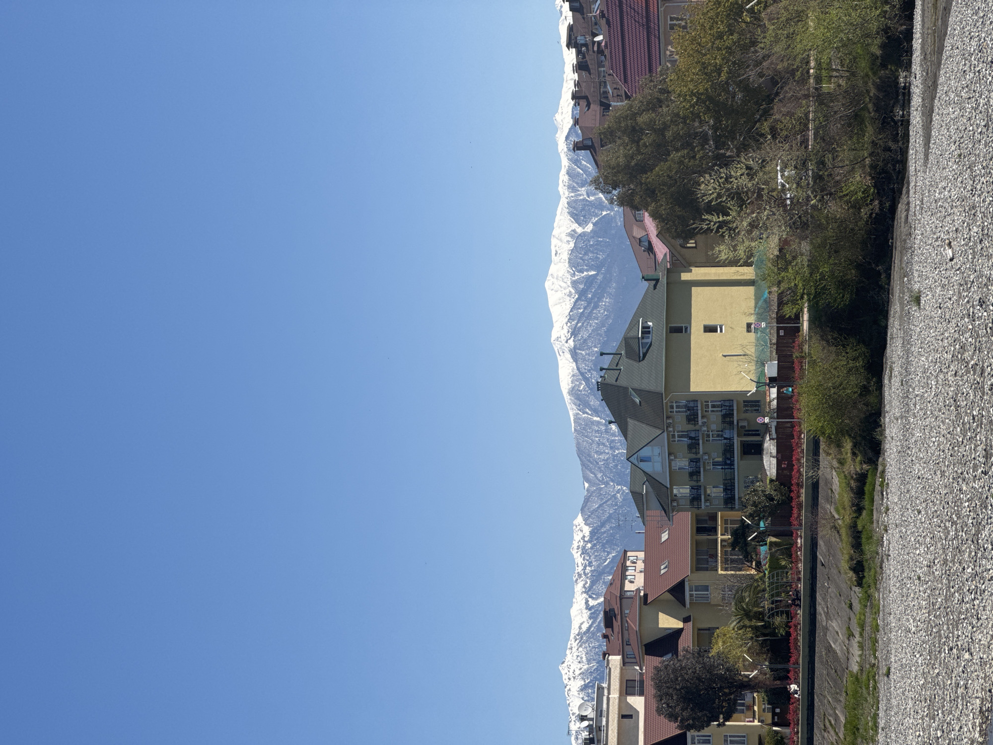 The image, when viewed upright, presents a picturesque landscape featuring a range of snow-capped mountains under a clear, bright blue sky. The mountains dominate the mid-ground, their white, rugged peaks creating a striking contrast against the expanse of sky. Below the mountain range, and extending into the foreground, is a collection of buildings, likely residential or mixed-use, displaying various architectural styles. Many have multiple windows, some with balconies or satellite dishes, and roof colors vary, including shades of red and green.
Interspersed among the buildings and along the foreground are numerous trees and bushes, indicating a lush environment. The immediate foreground consists of a wide, flat area covered in small stones or gravel, possibly a riverbed or a landscaped area, bordered by a concrete structure that might be a canal or riverbank.
The scene is illuminated by full daylight, suggesting a sunny day with excellent visibility. There are no people visible, and no overt activities are taking place, contributing to a tranquil and still atmosphere. A small, white sign with a red border and a diagonal line, resembling a prohibition sign, is faintly visible on a fence in the mid-ground among the buildings. The combination of urban development nestled against formidable mountains implies a location in a mountainous region, possibly a town or city near a large range.
