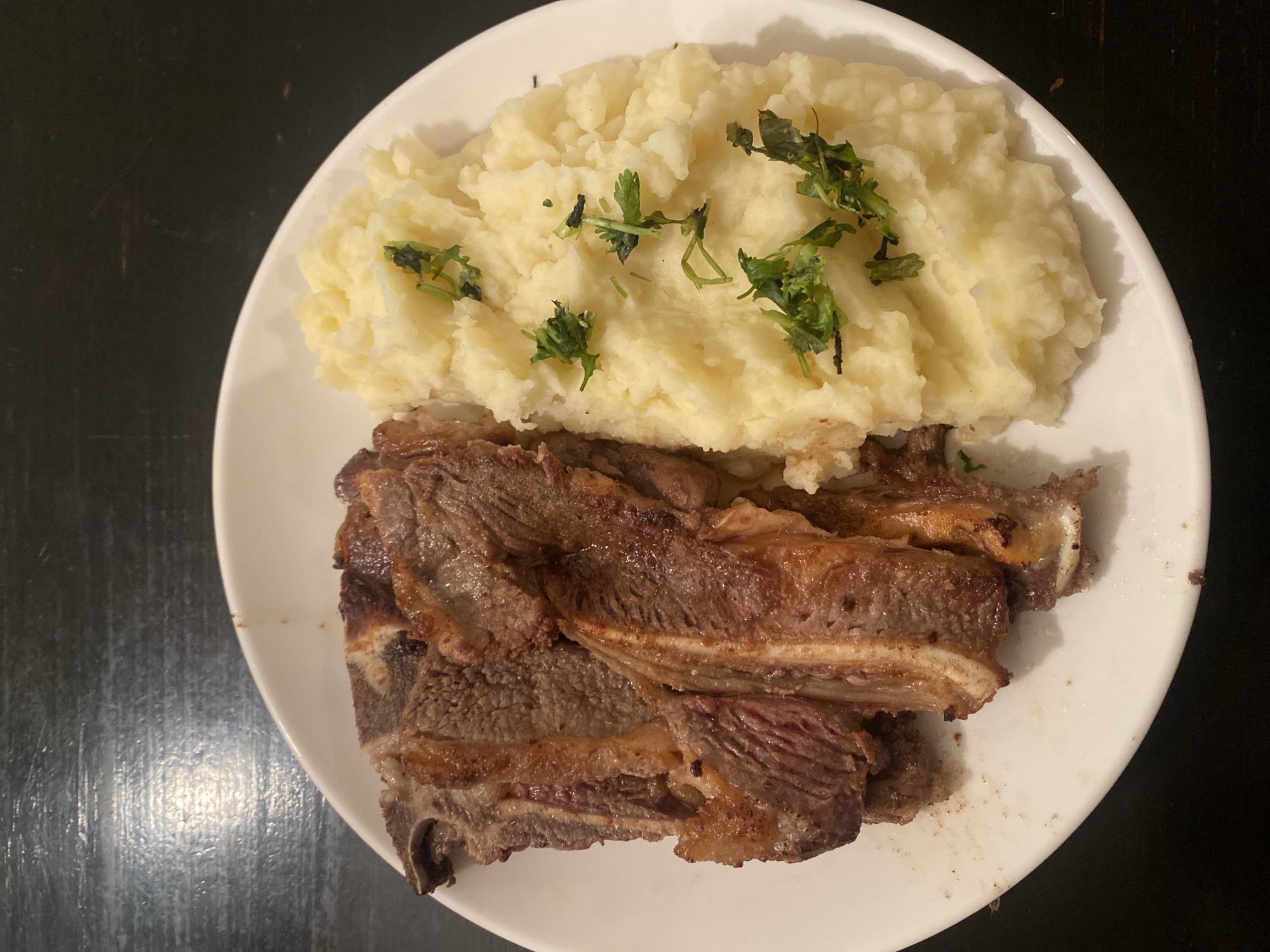 The image displays a prepared meal centered on a white, slightly oval plate. On the left side of the plate are approximately seven to eight segments of cooked meat, consistent with short ribs (asado de tira), exhibiting a dark brown, seared exterior with areas of visible fat and bone, while the interior flesh appears reddish-pink, indicating a medium-rare to medium doneness. To the right, a mound of light yellow, fluffy mashed potatoes occupies the other half of the plate. The mashed potatoes are garnished with scattered fresh green herbs, likely parsley or cilantro. The white plate rests on a dark, flat surface, presumably a table, which reflects ambient overhead lighting. Minor food residue is visible on the plate's left edge. No individuals, actions, or specific contextual elements are present in the frame. The image itself provides no visual indicators to identify its location within the city of Buenos Aires, Argentina.