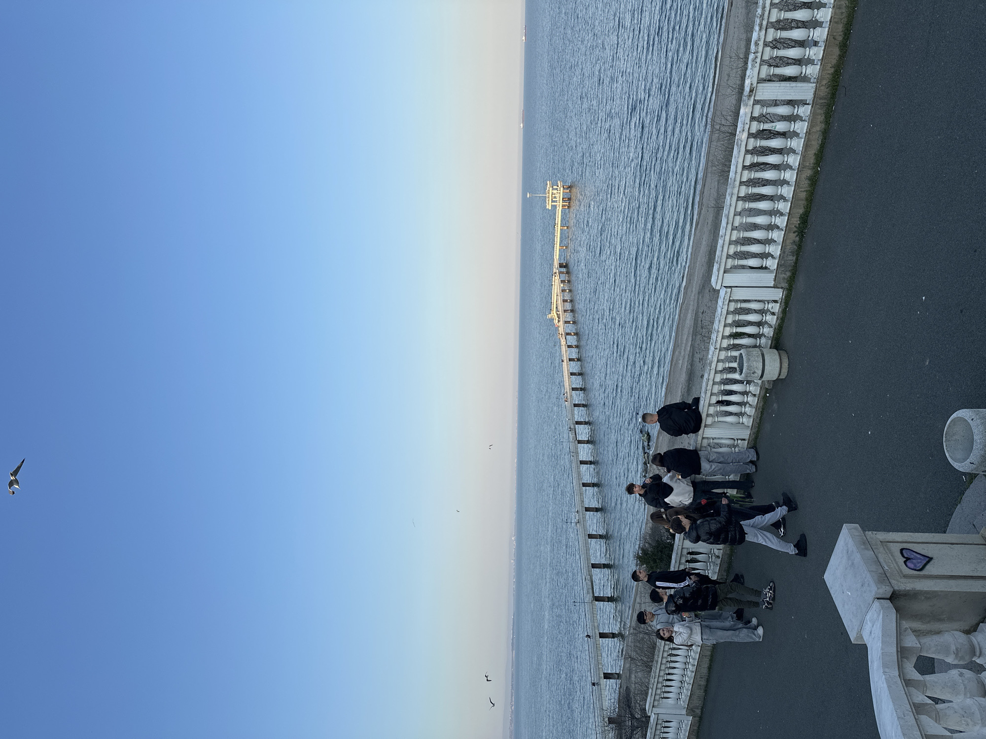The image depicts a group of approximately 10 people standing along a waterfront promenade in Burgas, Bulgaria. A pier stretches into the sea, with an observation deck at its end. The sky is clear, transitioning from blue to a lighter shade near the horizon. The promenade is bordered by a decorative white balustrade. People are dressed in casual clothing and some are wearing masks. A bird flies in the sky to the left. A gray heart is stenciled onto the side of the structure.
