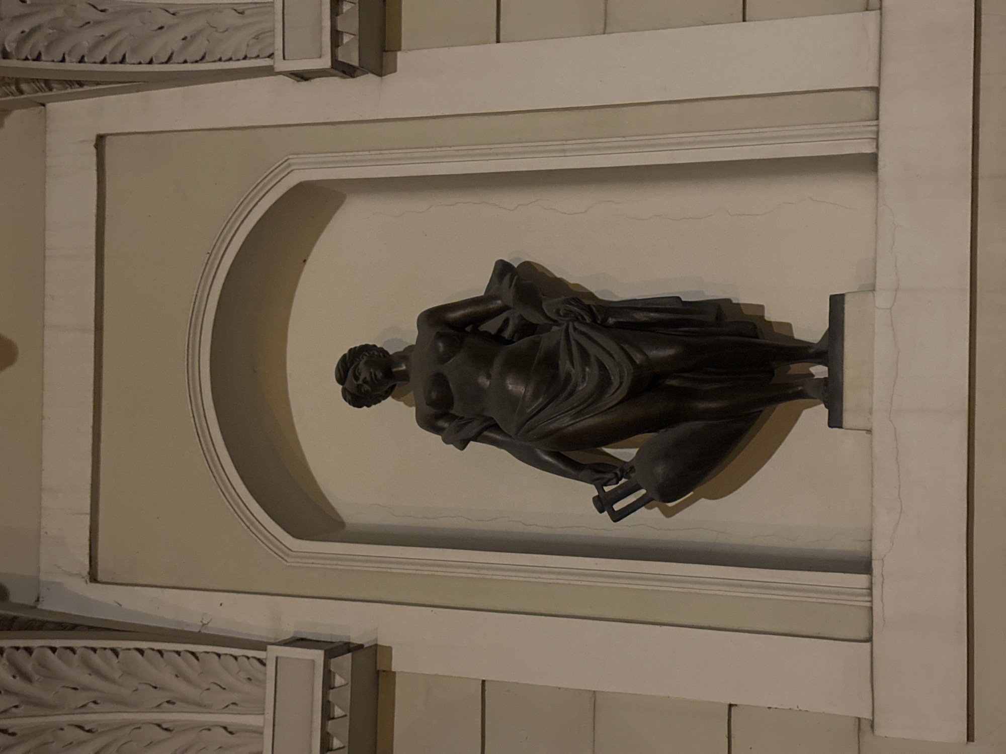 A bronze sculpture of a reclining female figure, draped with cloth, is set within an arched niche in a wall. The wall and the surrounding architectural elements are painted in shades of white and beige. The niche is framed by a simple white molding. The overall aesthetic is classical. The location is Riga, Latvia.