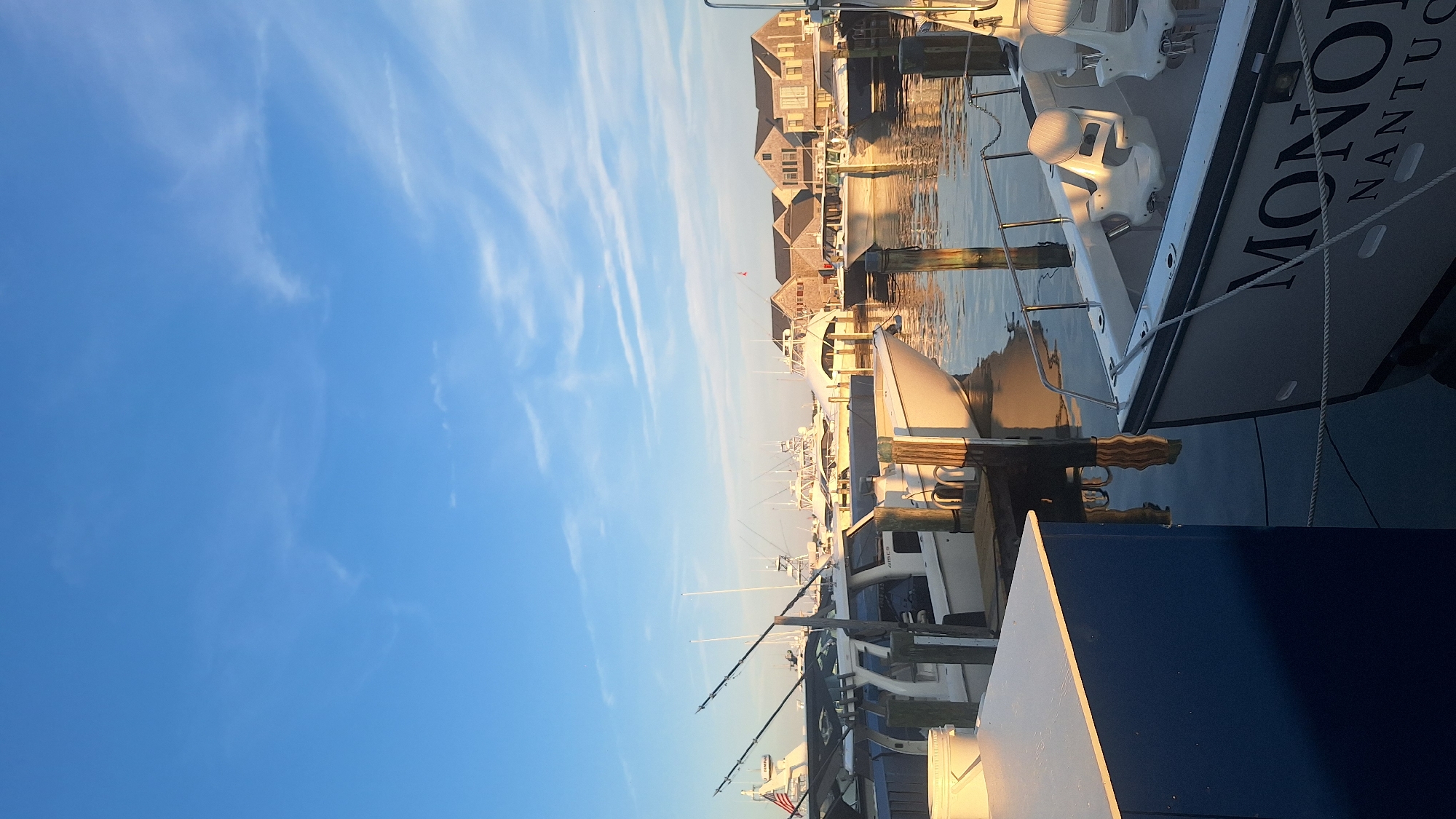 This is a vertical, eye-level, outdoor photograph of boats docked in a harbor in Nantucket, Massachusetts, United States of America, under a clear blue sky with thin, wispy clouds. In the foreground, the stern of a white boat with the name "MON" partially visible in large black lettering, followed by "NANTUCK" in smaller letters, is moored to a dock. The boat has white seats and stainless-steel railings. To the left of this boat is a blue-and-white structure.
Behind this foreground boat, other boats are docked in the harbor. Some have fishing poles attached. In the middle ground, a row of buildings with brown shingled roofs and wooden siding lines the waterfront. These buildings appear to be shops or residences. The water in the harbor reflects the sky and the boats. The lighting suggests it is either early morning or late afternoon, with the sun casting a warm glow on the scene.