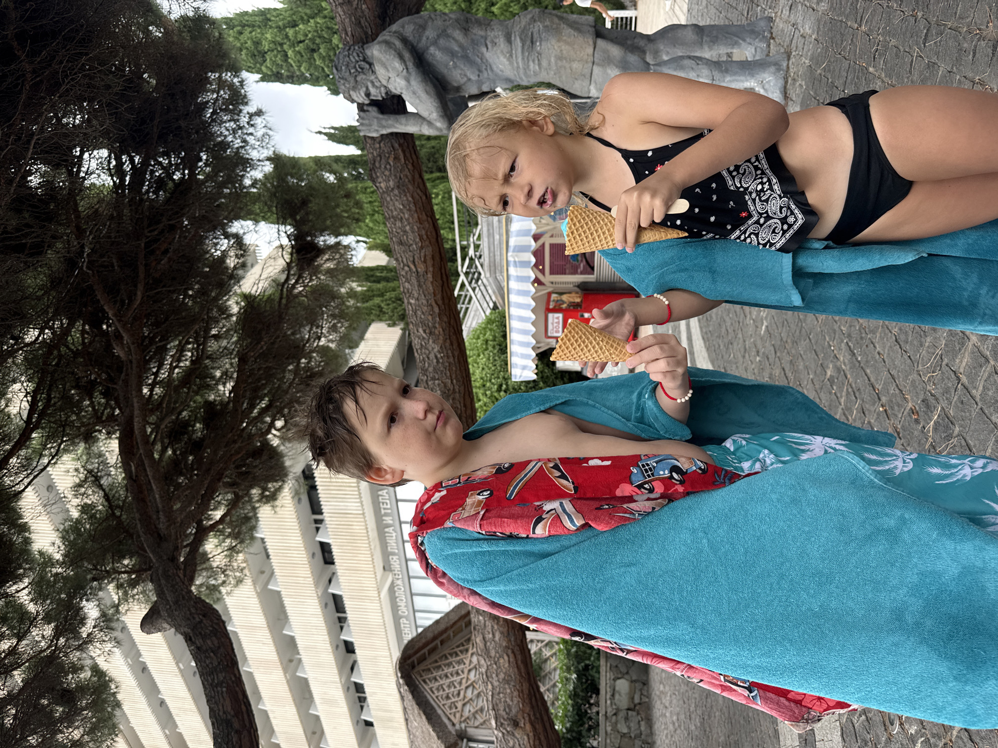 Two children, a boy and a girl, stand outside a building in Yalta, Ukraine. The boy, wrapped in a turquoise towel over a red and white patterned garment, holds an ice cream cone. The girl, wearing a black swimsuit and a turquoise towel, also holds an ice cream cone and appears to be speaking. Behind them, a large tree branch extends across the frame. A building with multiple balconies is visible in the background, along with trees and a statue of a man. The ground is paved with cobblestones.
