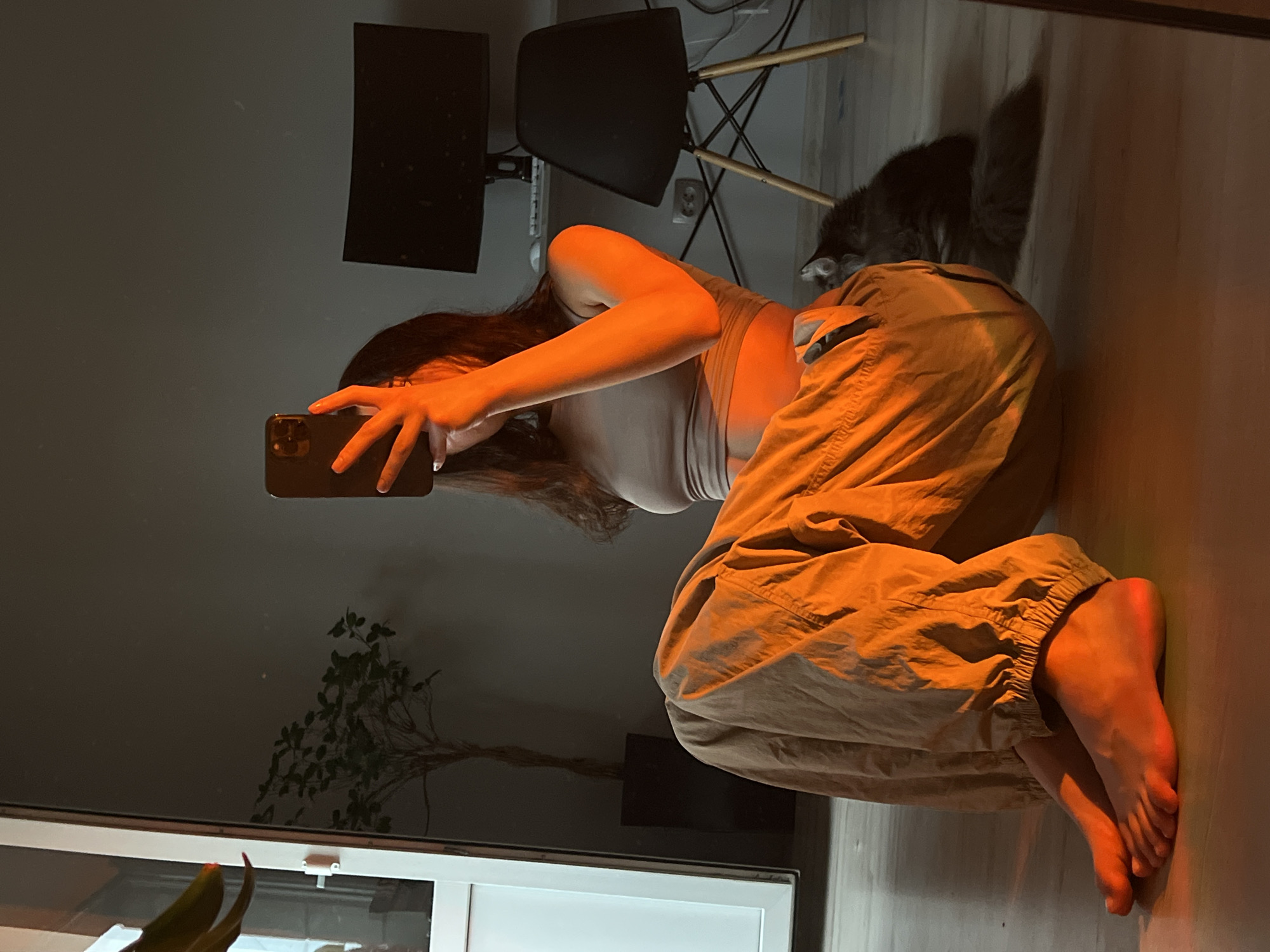 The image, taken in Ternopil, Ukraine, depicts a person kneeling on a floor, holding a smartphone to take a selfie. The individual has long, dark hair and is wearing a light-colored crop top and loose-fitting tan pants. The setting appears to be an indoor space, possibly a home, with warm, artificial lighting casting an orange glow on the scene.
In the background, a gray cat is visible, partially obscured by the person's legs and the backdrop. There are also glimpses of furniture, including a black chair with wooden legs and what appear to be computer monitors mounted on a wall. A plant with thin branches is situated to the left of the person. The floor is reflective, suggesting it might be polished wood or tile, and it catches the warm lighting. The overall mood is intimate and casual, with the warm lighting creating a cozy atmosphere. The image is oriented vertically, and it's impossible to determine the specific time of day or weather conditions outside. No visible text is present in the image.