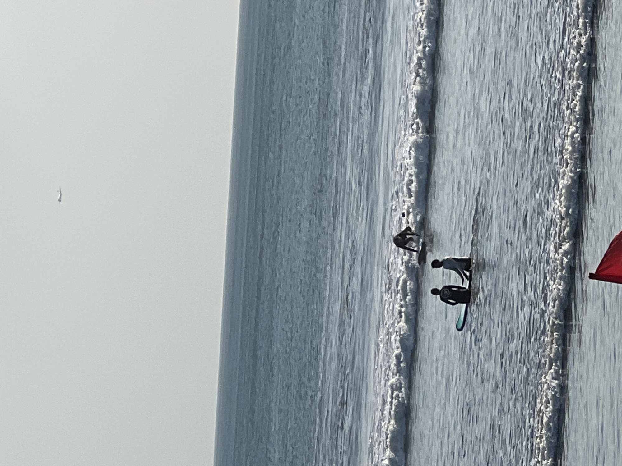 The image depicts a serene ocean scene with several individuals engaged in surfing activities in the waters of Kuta, Bali, Indonesia. The ocean exhibits gentle waves with white foam crests, indicating calm conditions. In the foreground, two figures, appearing to be surfers, stand waist-deep in the water, each with a surfboard. One figure is positioned in front of the other and is partially obscured by the waves. Further out, another individual is seen kneeling on their surfboard, preparing to ride a wave. In the upper portion of the image, a distant airplane is visible against a hazy, light-colored sky, suggesting a clear day. The overall ambiance of the scene is tranquil and recreational, characteristic of a beach environment.