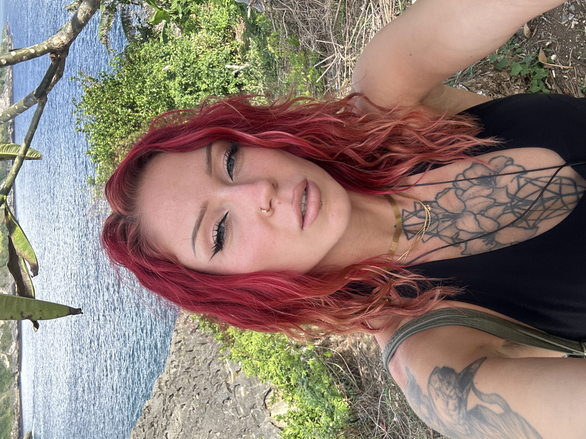 A woman with red hair is taking a selfie outdoors. She is wearing a black top and has tattoos on her chest and arm. The background includes the ocean, vegetation, and a rocky cliff. Based on the environment depicted, this could be in the Nusa Penida area of Indonesia.