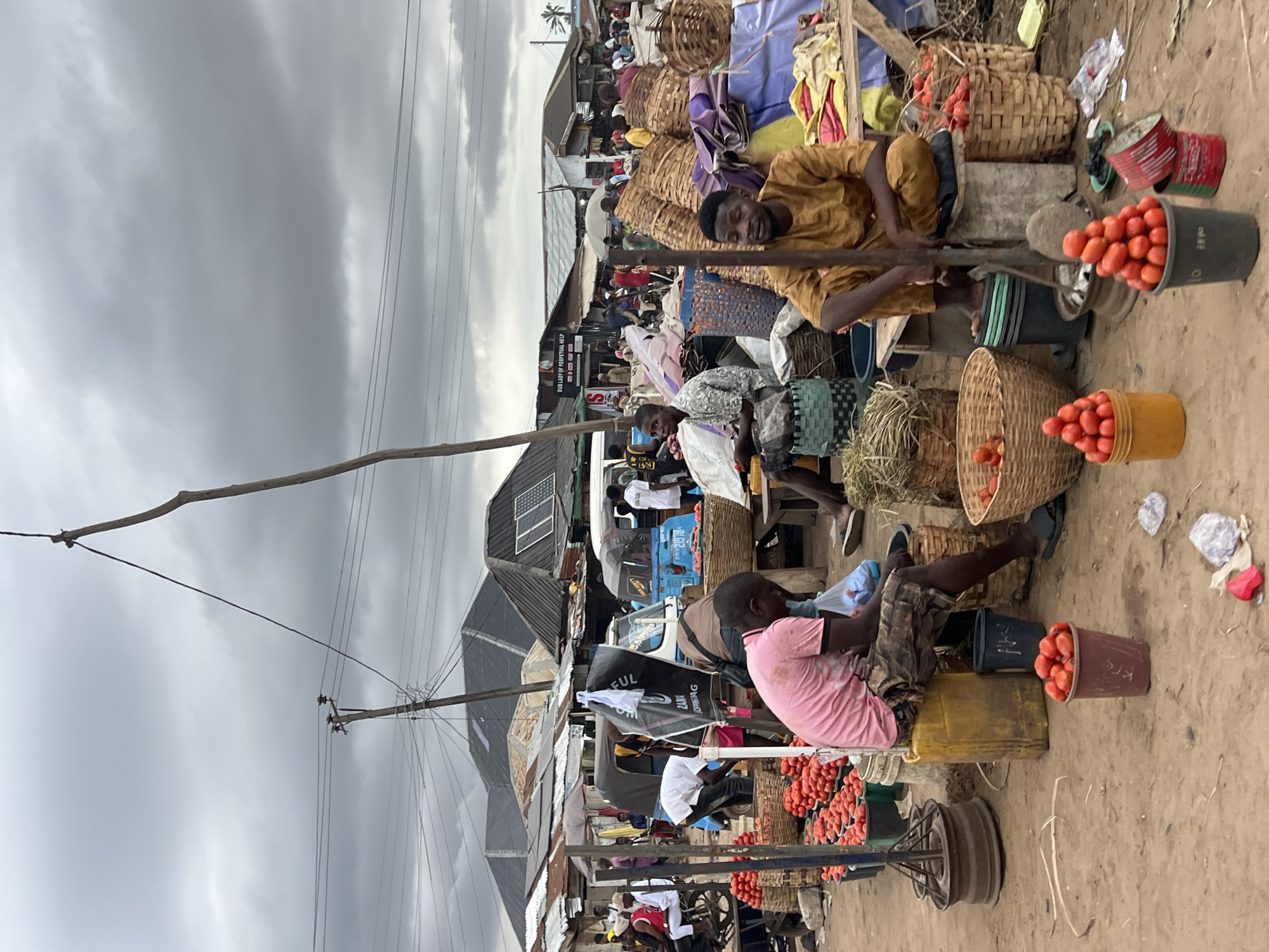 This image depicts an outdoor market scene in Oko Amakom, Nigeria. The sky is overcast with gray clouds. In the foreground, two men are seated. The man on the left wears a pink t-shirt and patterned shorts, sitting on a yellow container. The man on the right wears a mustard-colored traditional garment and is seated on a wooden stool. Both men appear to be vendors.
Numerous baskets and buckets filled with red tomatoes are visible throughout the scene, indicating a produce market. Several larger woven baskets are stacked, some appearing empty, others with produce or fabric. In the background, various market stalls with corrugated metal roofs and exposed wooden structures are present. Several auto-rickshaws, known locally as Keke-napep, are parked. Some market-goers are visible in the middle ground, engaged in activity. Power lines and utility poles are present in the upper portion of the image. The ground is dirt and appears worn.