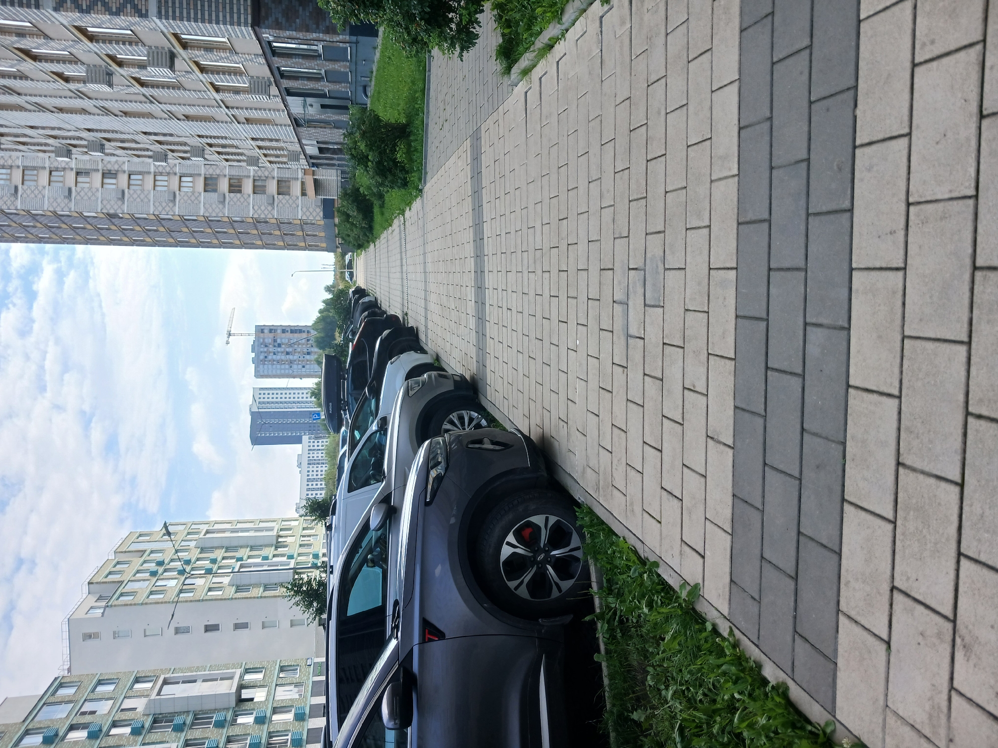 The image depicts a street view in a modern urban development in Yekaterinburg, Russia. A row of parked cars, predominantly SUVs and sedans, lines a paved walkway made of interlocking concrete pavers in shades of gray and beige. The cars are parked parallel to the walkway.
To the right of the walkway, a modern apartment building with a brick facade and numerous windows rises several stories high. Green grass and small bushes are visible along the base of the building.
To the left of the walkway, across from the brick building, is another apartment complex composed of lighter-colored buildings with blue architectural accents. These buildings are also multi-story structures. In the distance, more buildings and a construction crane are visible, suggesting ongoing development in the area. The sky is partially cloudy.
The scene is tranquil, with no people or specific activities evident. The overall impression is one of a residential area in a contemporary Russian city.