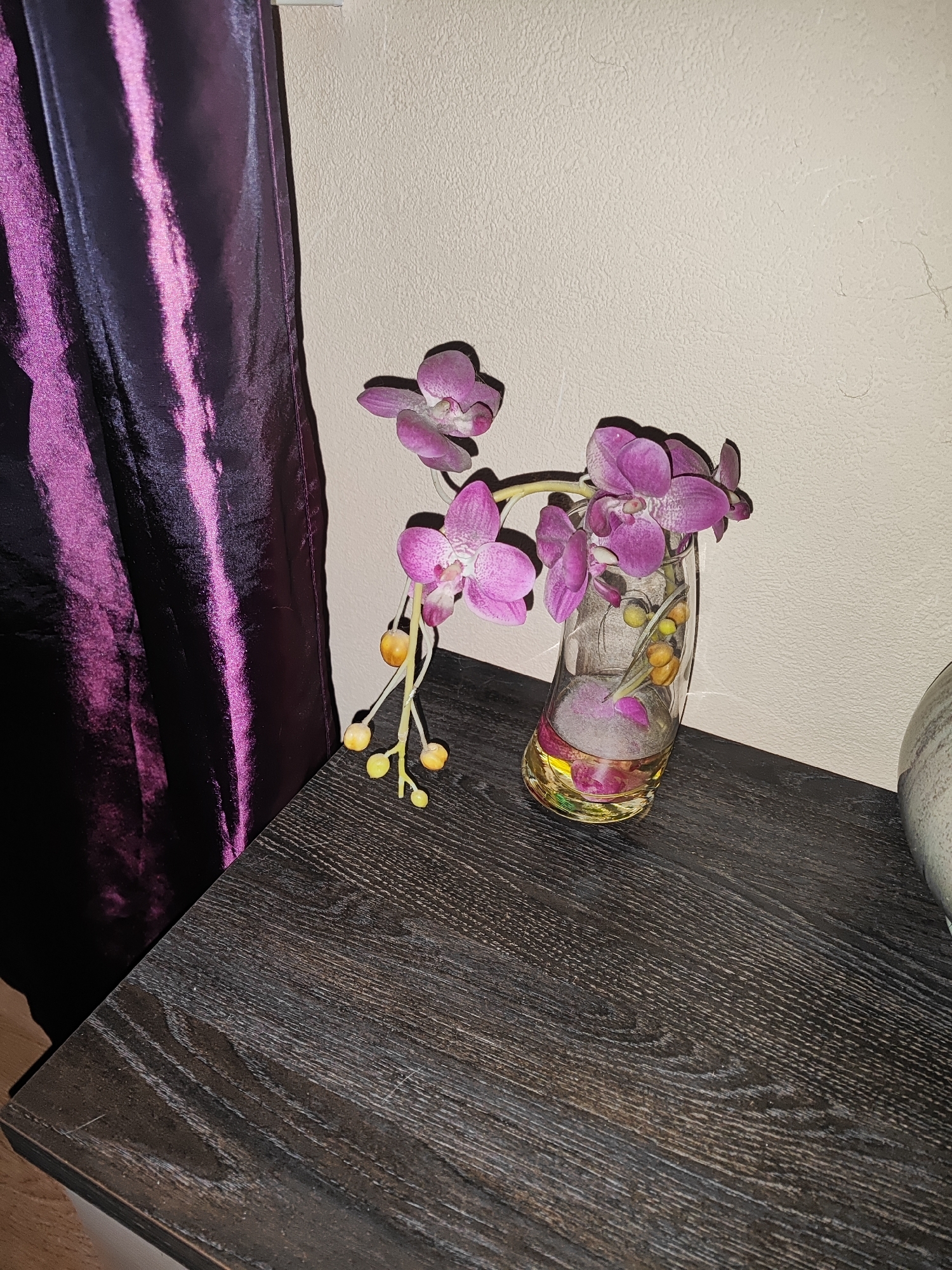 A still life composition on a dark wood surface features a glass vase containing artificial purple orchids with yellow accents. The vase is filled with water and decorative stones. To the left, a dark purple fabric with vertical pink stripes, likely a curtain, is draped. The background is a textured, light-colored wall. The scene is indoors, likely in a domestic setting. The location is Gvardeysk, Russia.