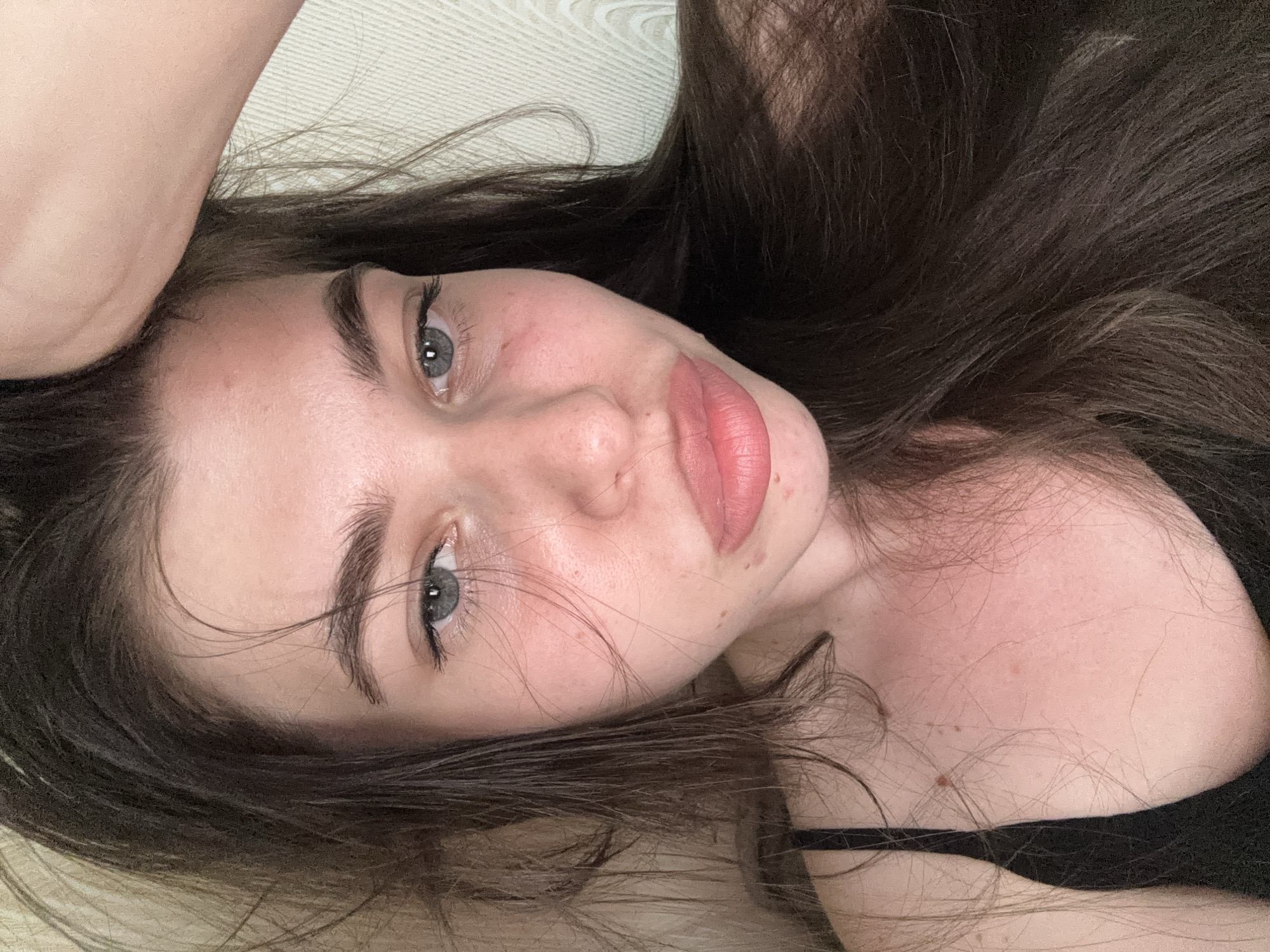 A close-up, selfie-style shot of a young woman lying on her back. She has fair skin, dark brown hair, and light-colored eyes. Her lips are full and pink. She is wearing a black top. The background is a light-colored surface with a subtle textured pattern. No location of Мещёры, Russia is visible in this image.